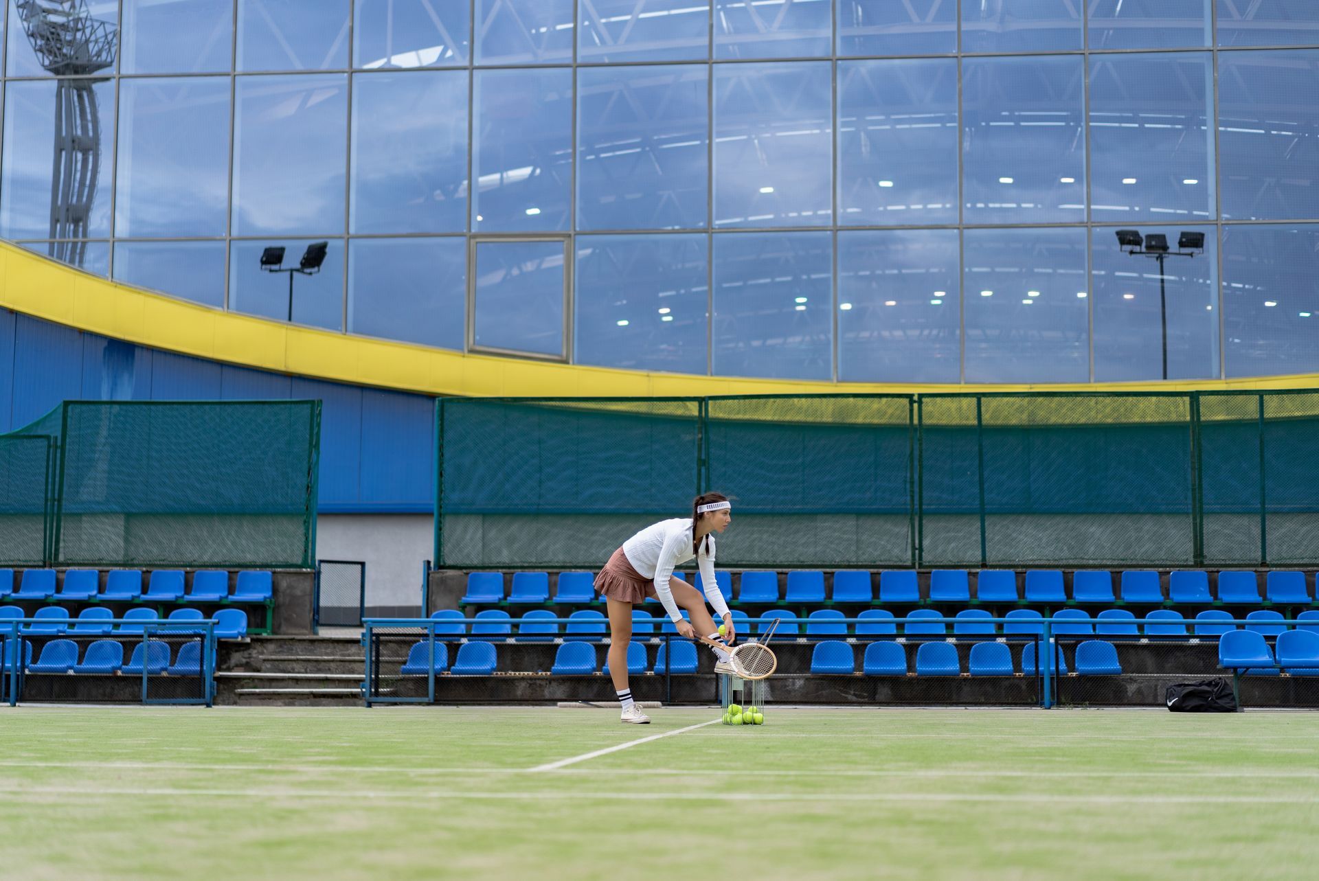 Woman on tennis court bends to pick up balls, bleachers and stadium in background.