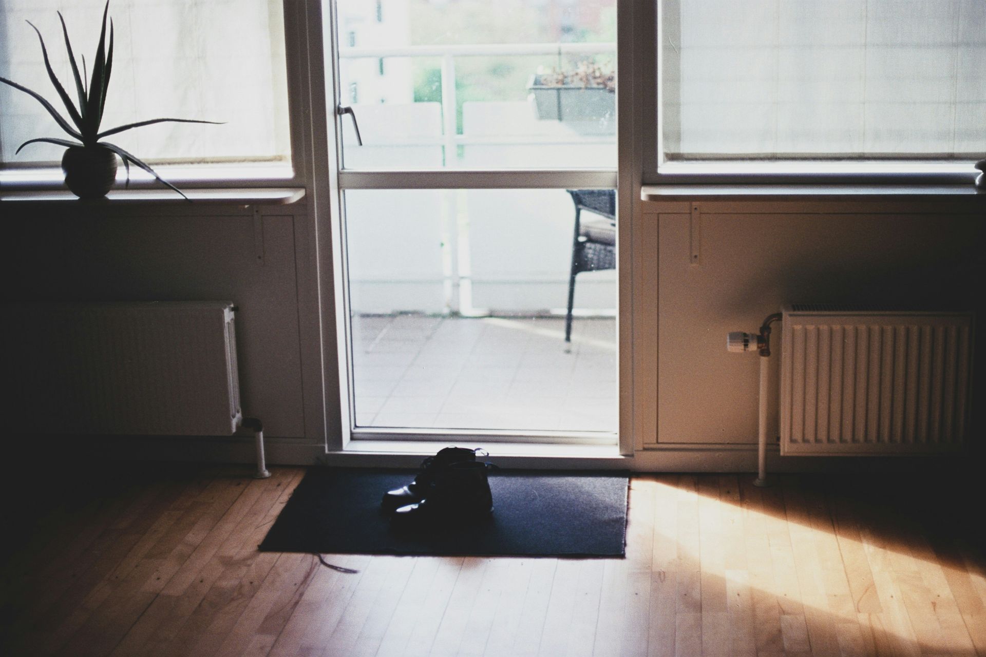 Open doorway to a balcony; a plant, shoes on mat, and radiator inside; sunlight streams in.