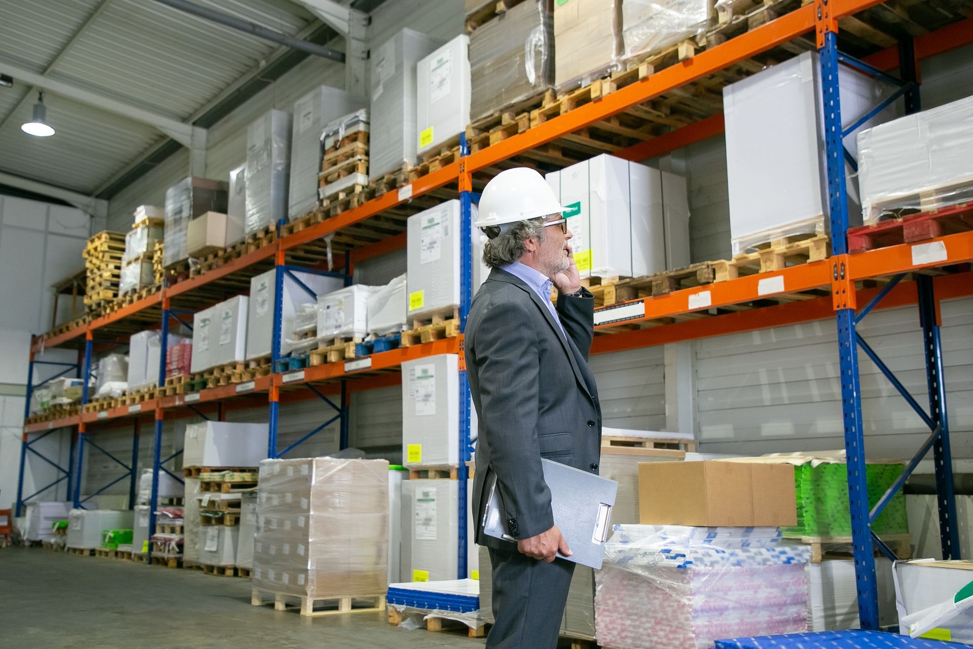 A warehouse worker in a yellow forklift moves a pallet of stacked items near a conveyor belt.