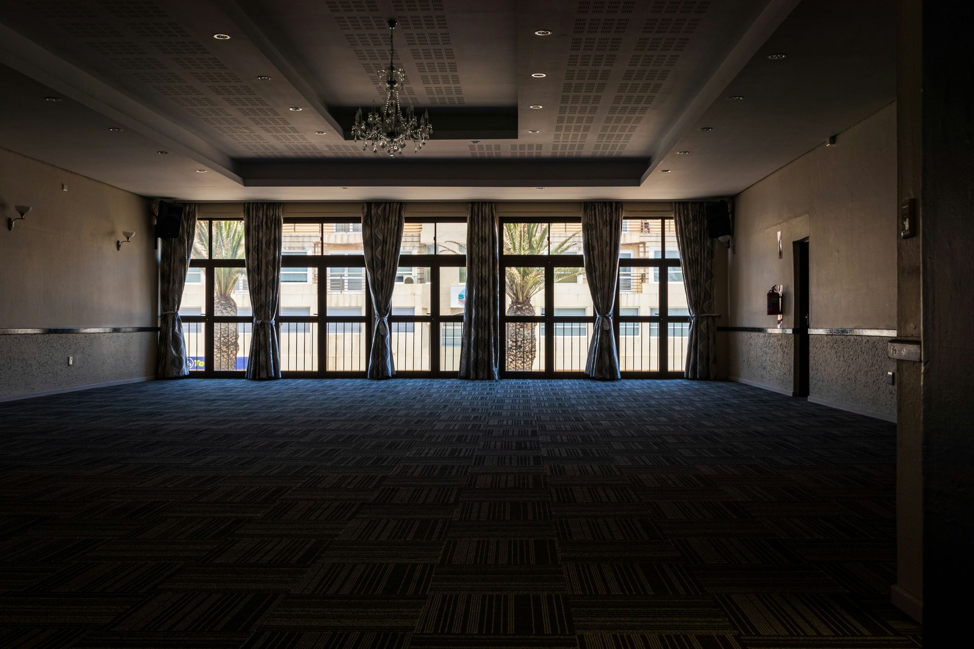Empty ballroom with large windows, dark carpet, and hanging chandelier.