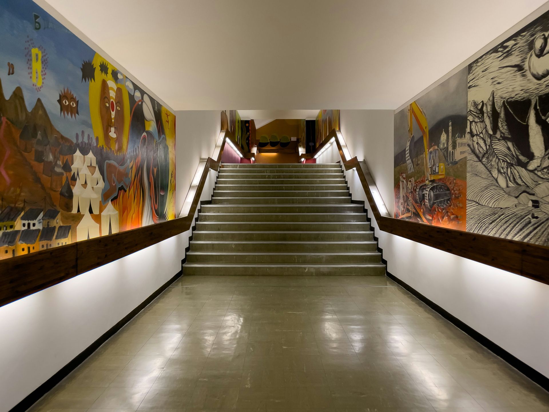 A long hallway with stairs, flanked by murals and illuminated by strip lighting, leading to an opening.