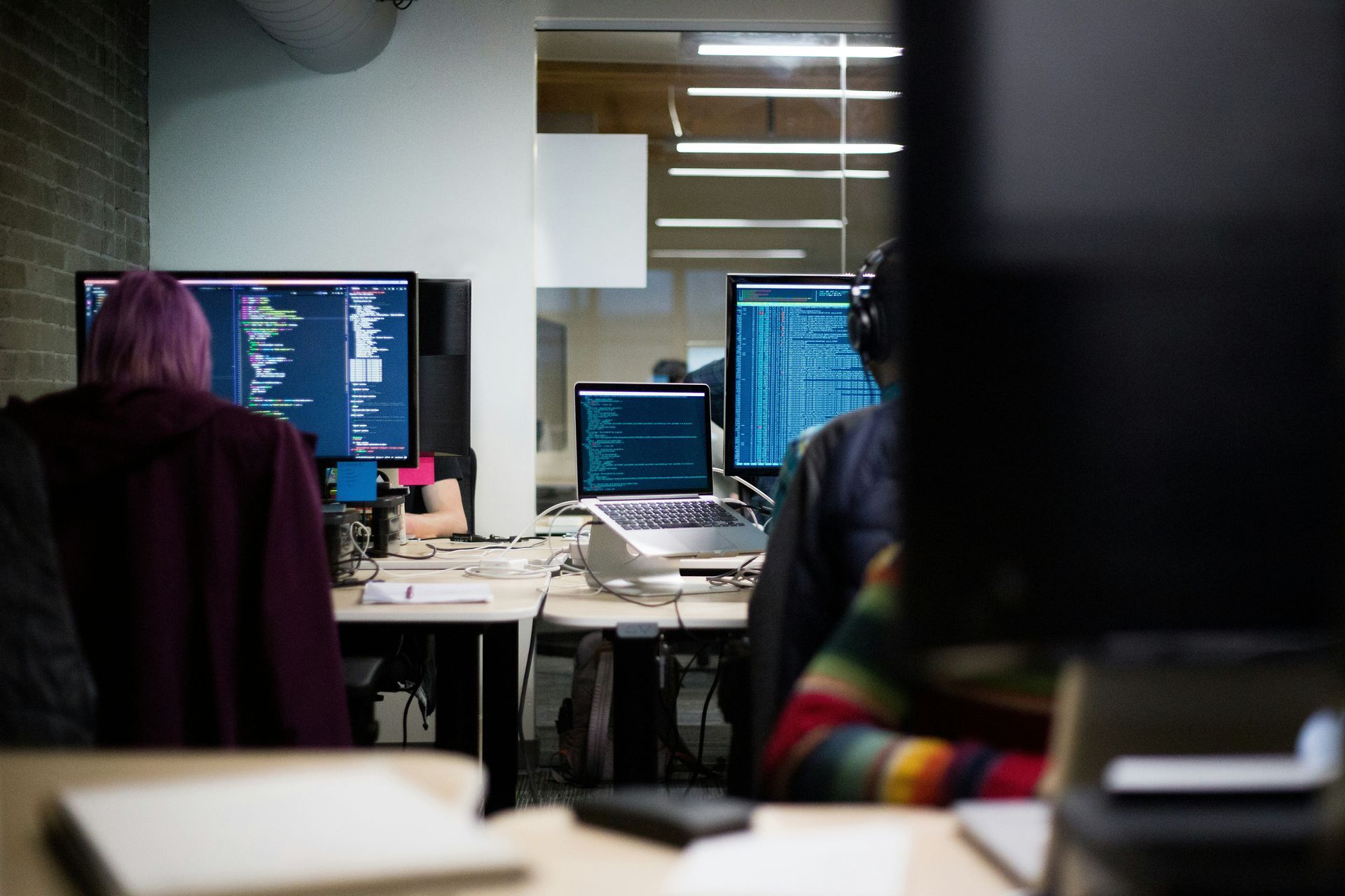 People coding at their desks in a modern office. One with purple hair. Multiple computer monitors.
