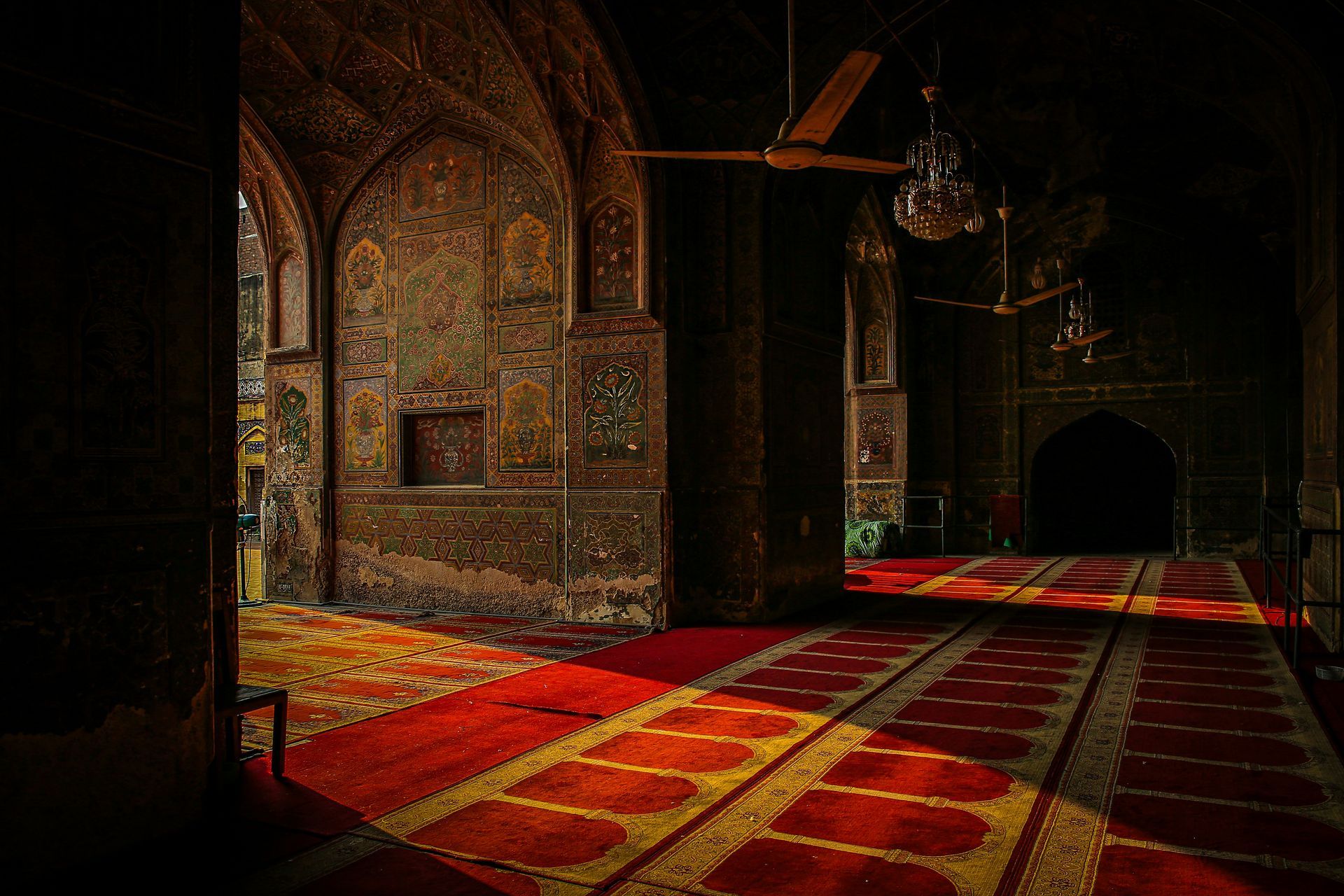Inside a mosque with ornate walls and red carpets, illuminated by sunlight.