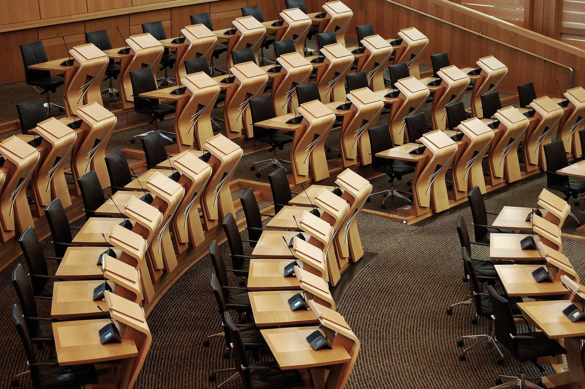 Rows of empty wooden desks and chairs in a formal, wood-paneled hearing room.