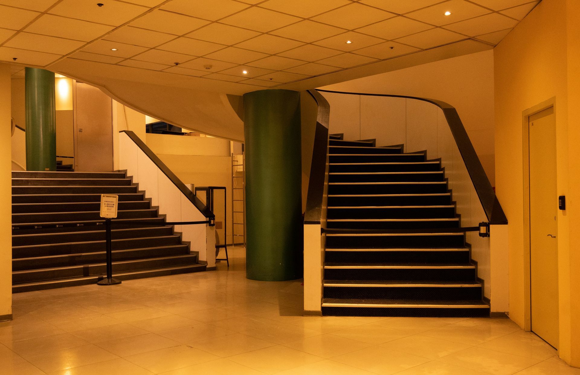 Staircase in building interior. Two staircases with black steps flank green pillar. Yellow walls and ceiling.
