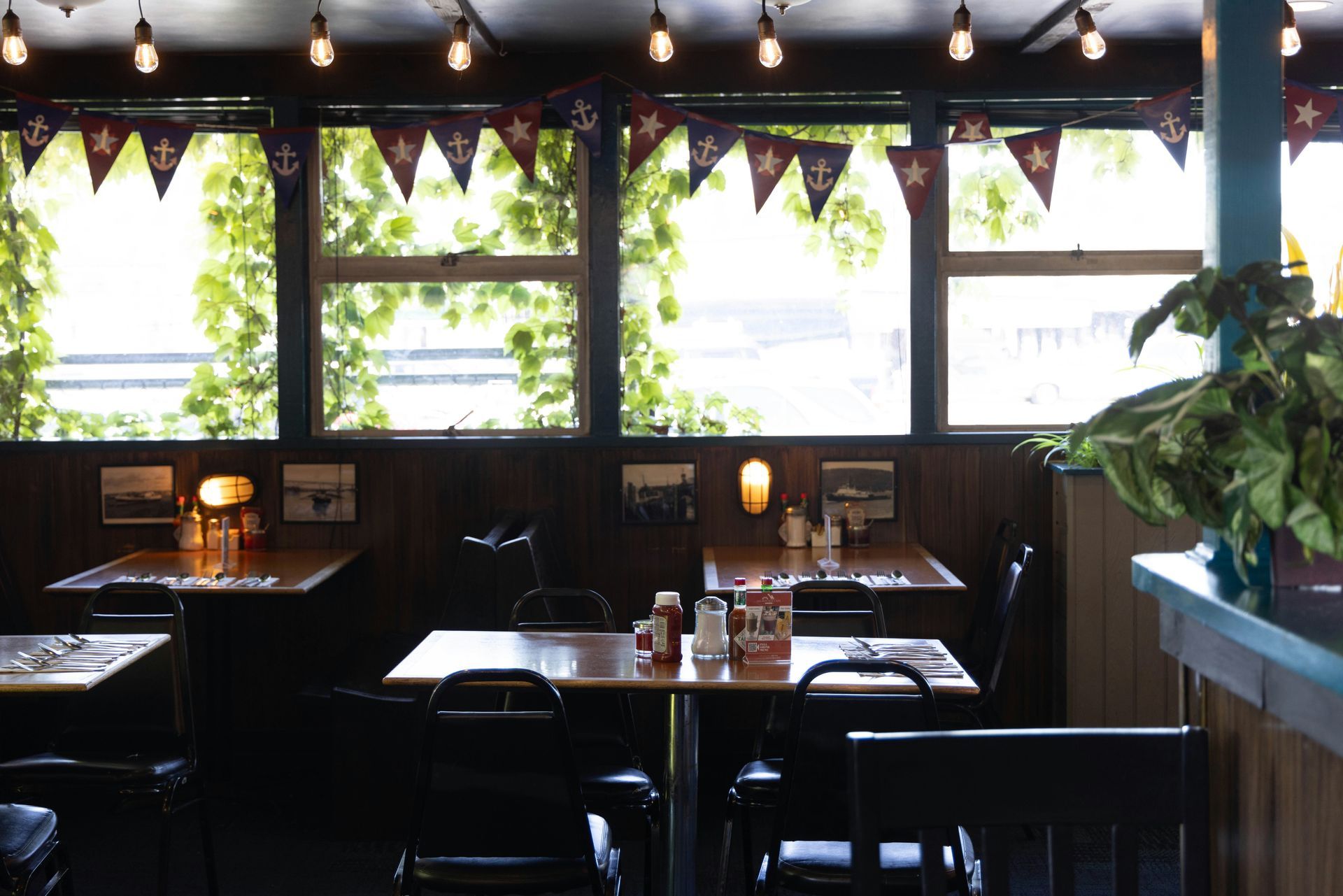 Interior of a restaurant with square tables, dark chairs, and nautical-themed decorations near windows with greenery.
