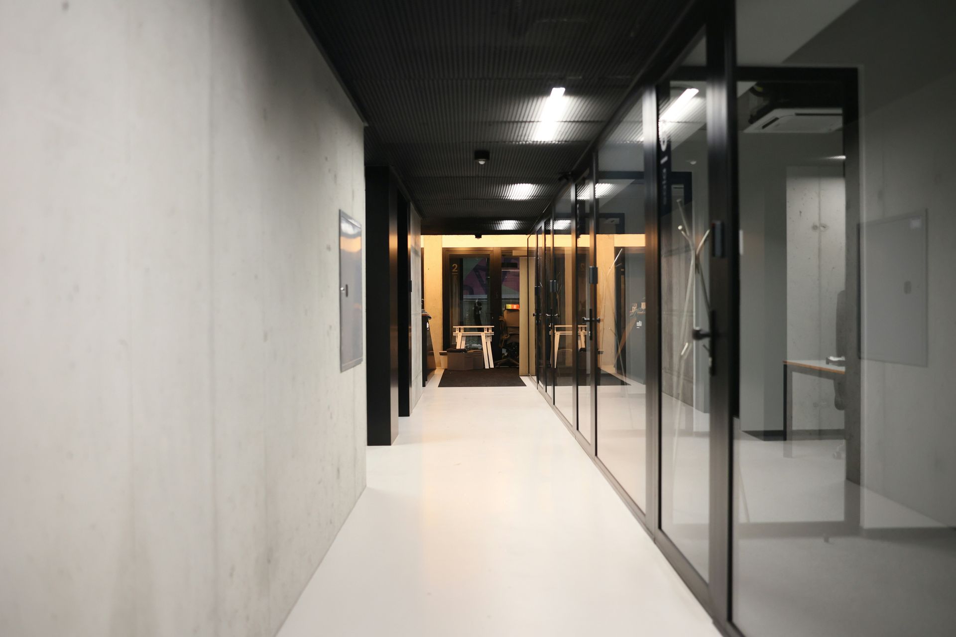 Hallway with glass-walled offices on the right, a concrete wall on the left, and a light-filled doorway at the end.