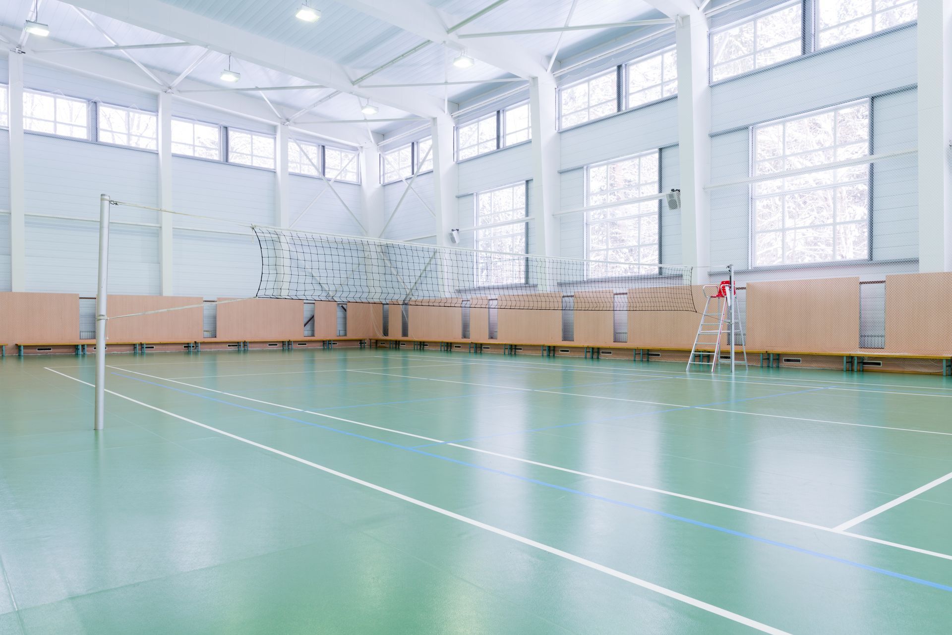 Empty volleyball court, green floor, white lines, net, wooden panels along the sides, bright interior.