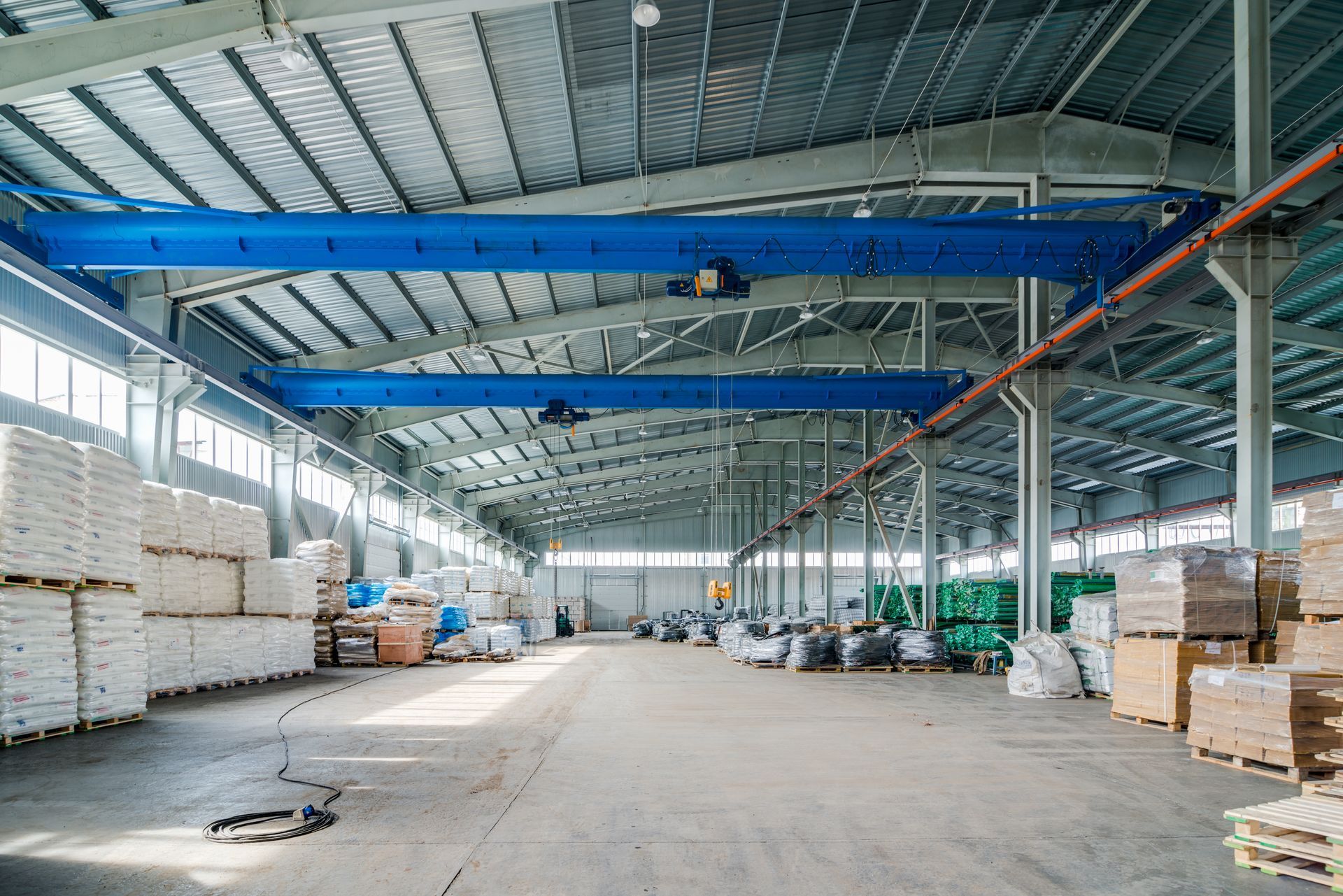 Warehouse interior with a blue overhead crane, goods stacked, and sunlight.