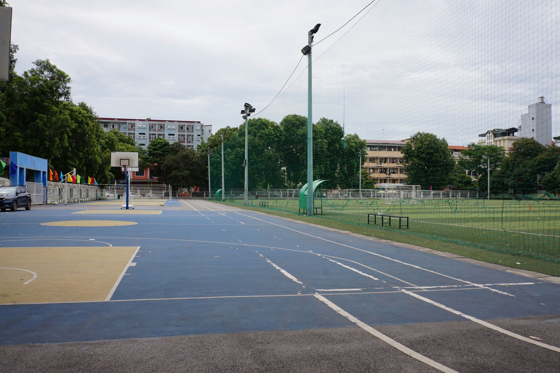 Outdoor sports court with basketball hoop, track, and soccer field under a cloudy sky.