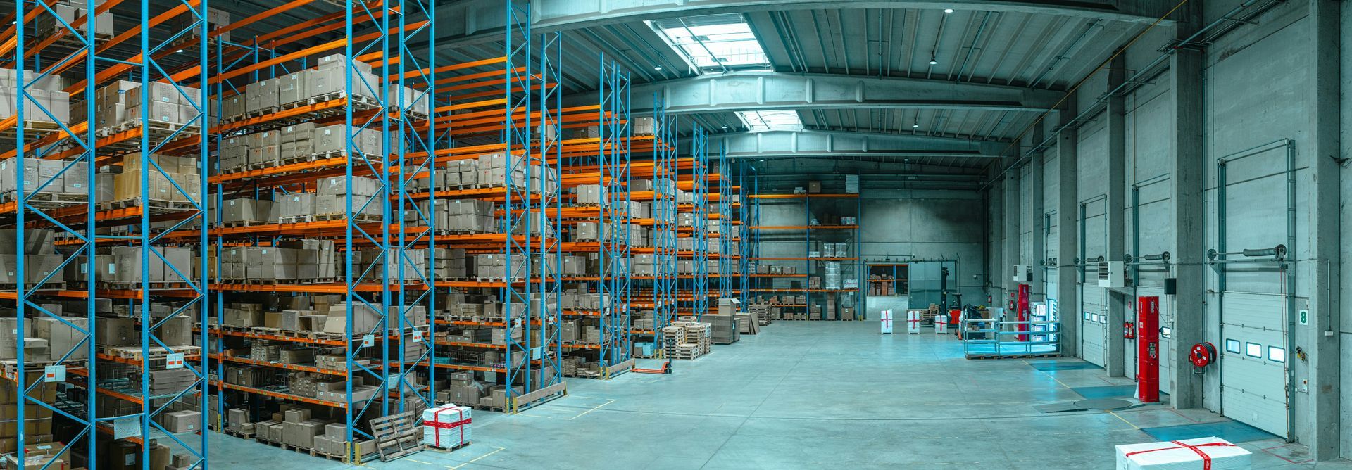 Warehouse interior with high shelves stacked with boxes. Industrial setting with loading doors and equipment.