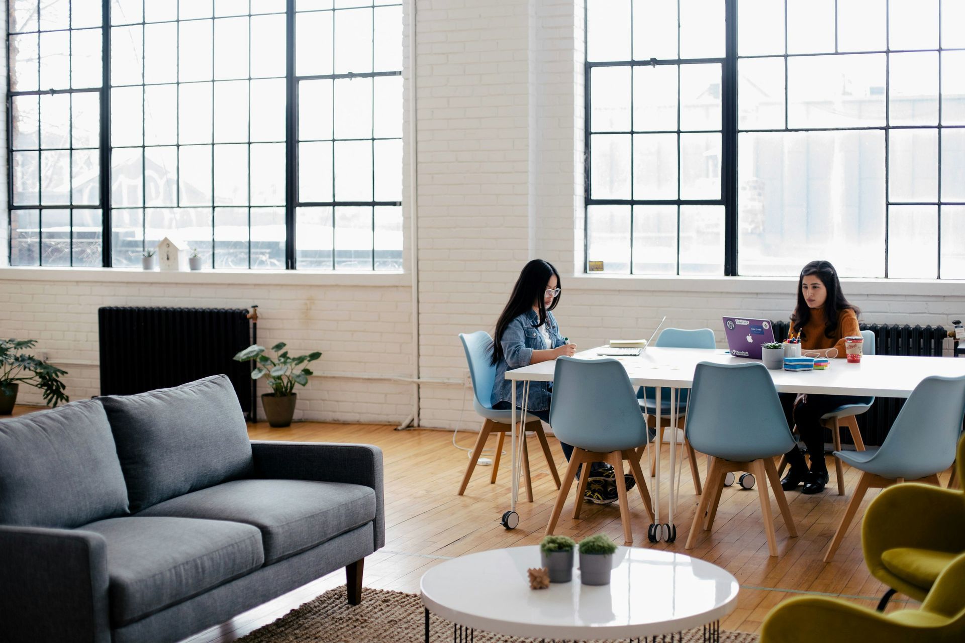 Two women at a table in a bright office. Grey sofa, round table, and big windows.