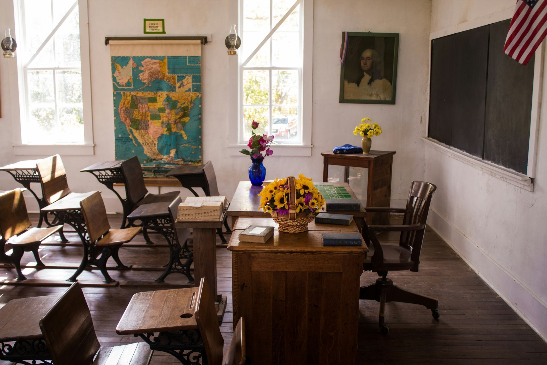 Old-fashioned classroom: desks, blackboard, map, teacher's desk with flowers. American flag. Bright windows, wooden floors.