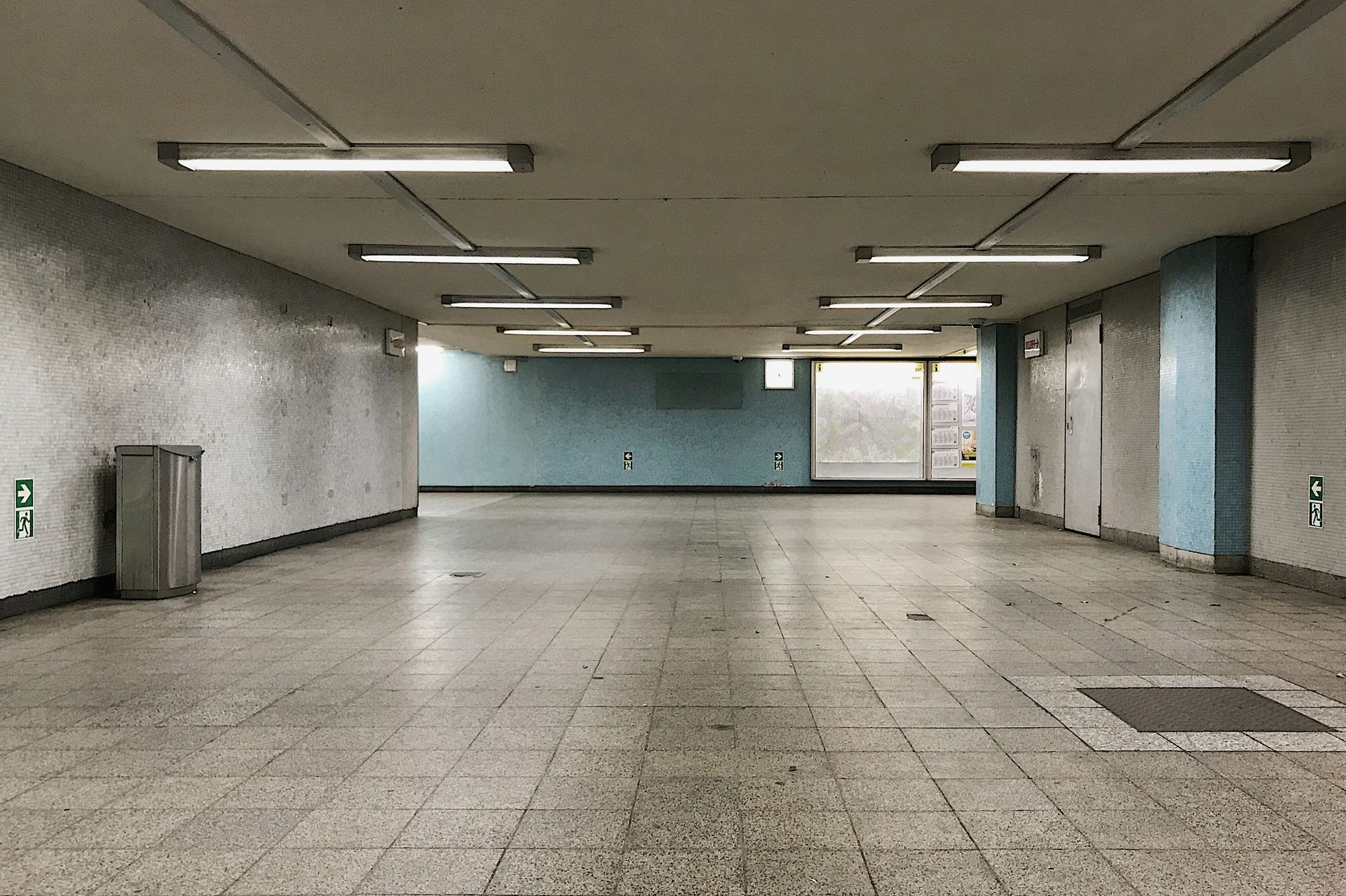 Empty, tiled subway tunnel with fluorescent lights. A trash can sits on the left, doors on the right.