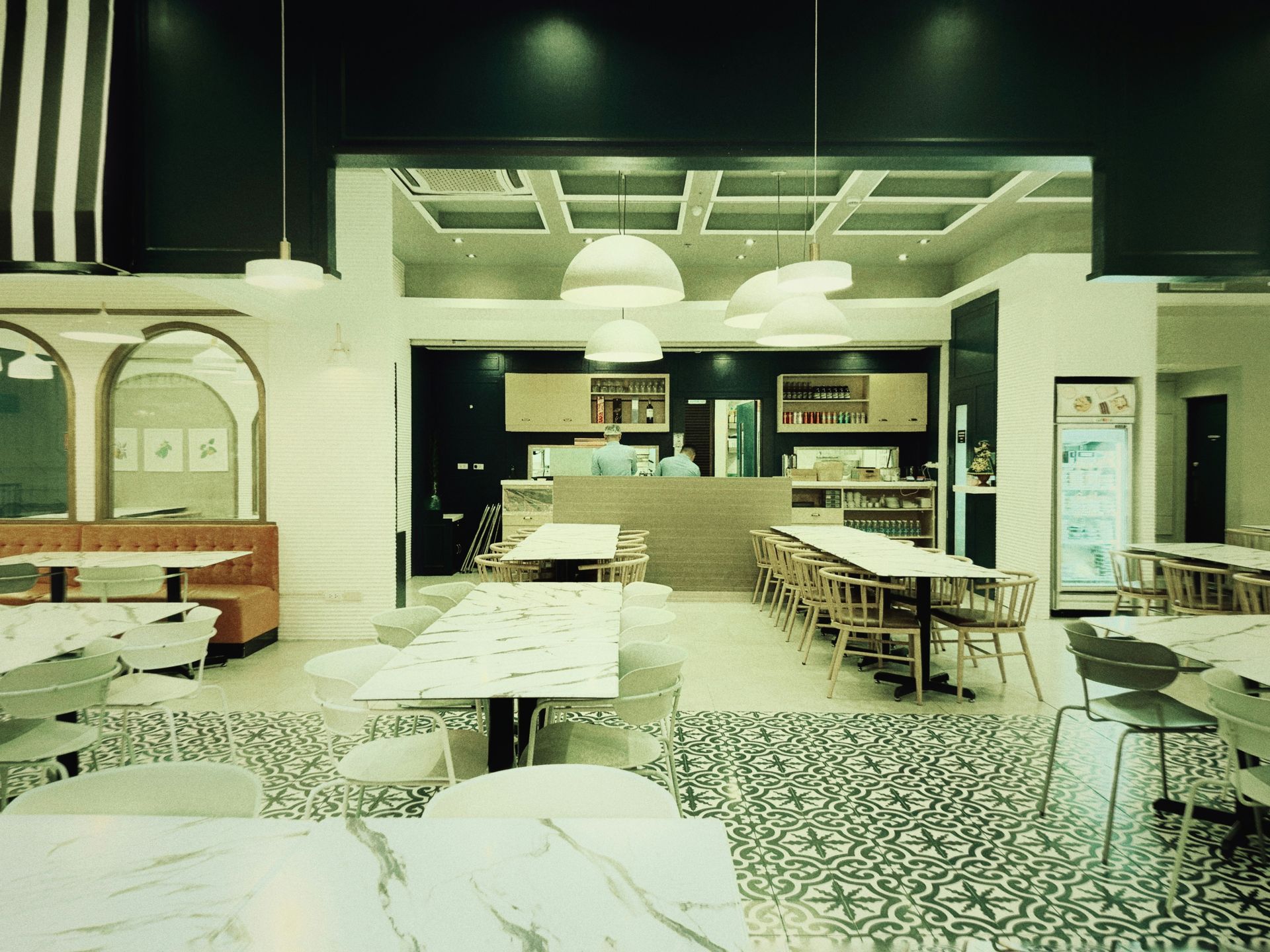 Interior of a modern restaurant: white tables, wood chairs, marble floor, bar, and black accent wall.