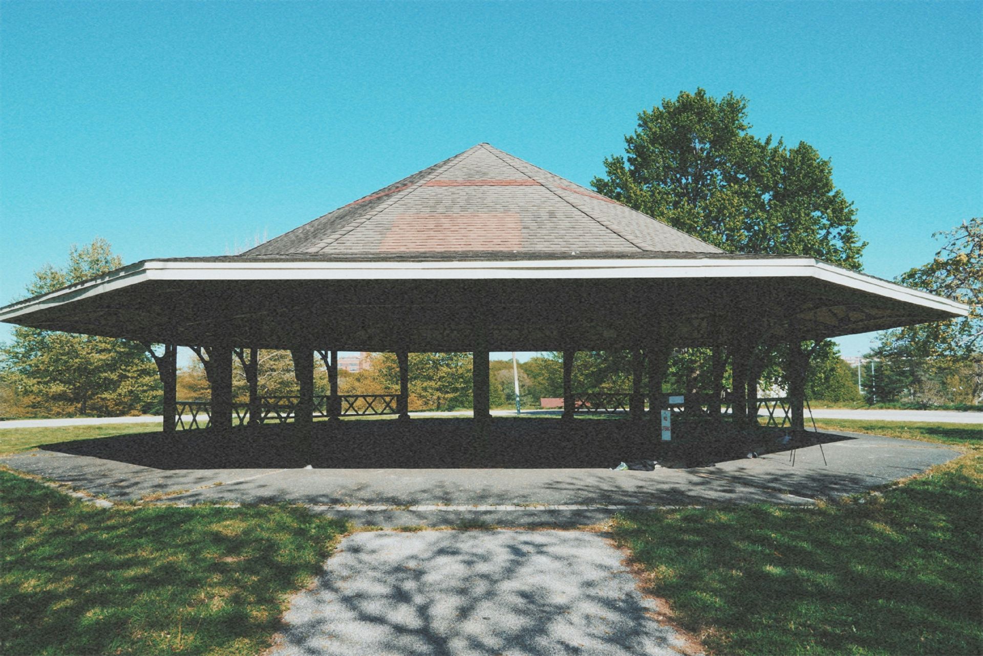 Octagonal pavilion with brown roof, open sides, on a paved area in a park, sunny day.