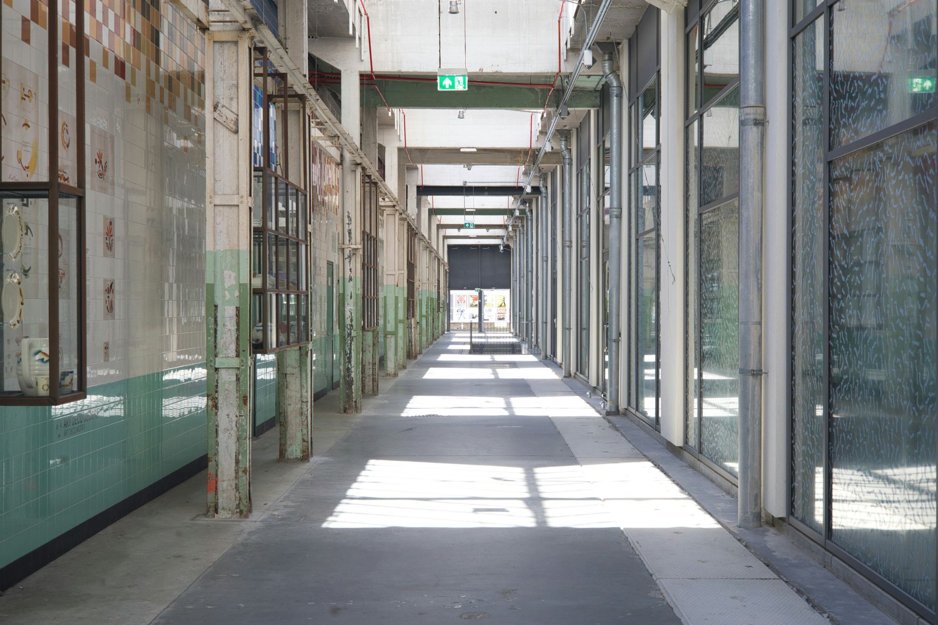 Long hallway of industrial building with glass windows on right, worn walls and columns, bright sunlight.