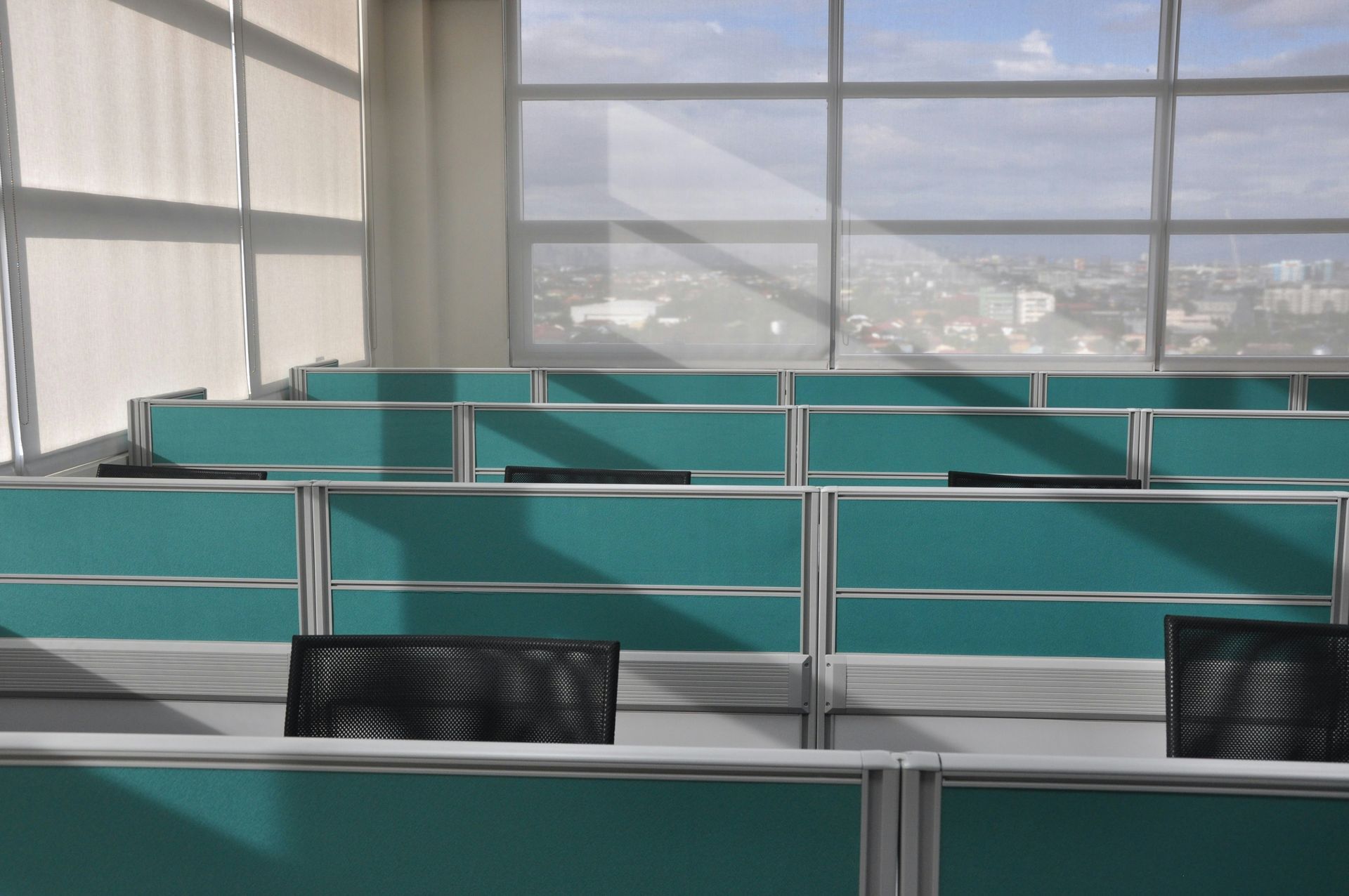 Empty office cubicles with teal dividers, black chairs, and large windows overlooking a cityscape.