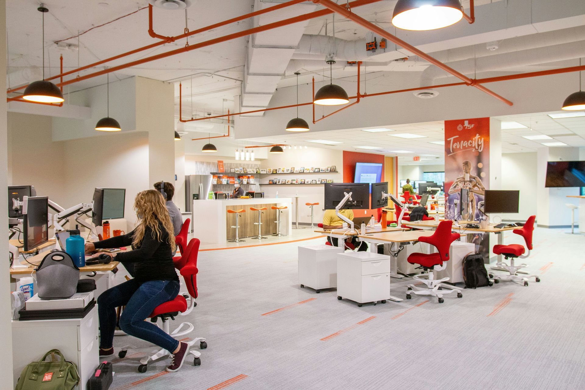 Open-plan office with people working at computers. Red chairs, white desks, and exposed ceiling pipes.