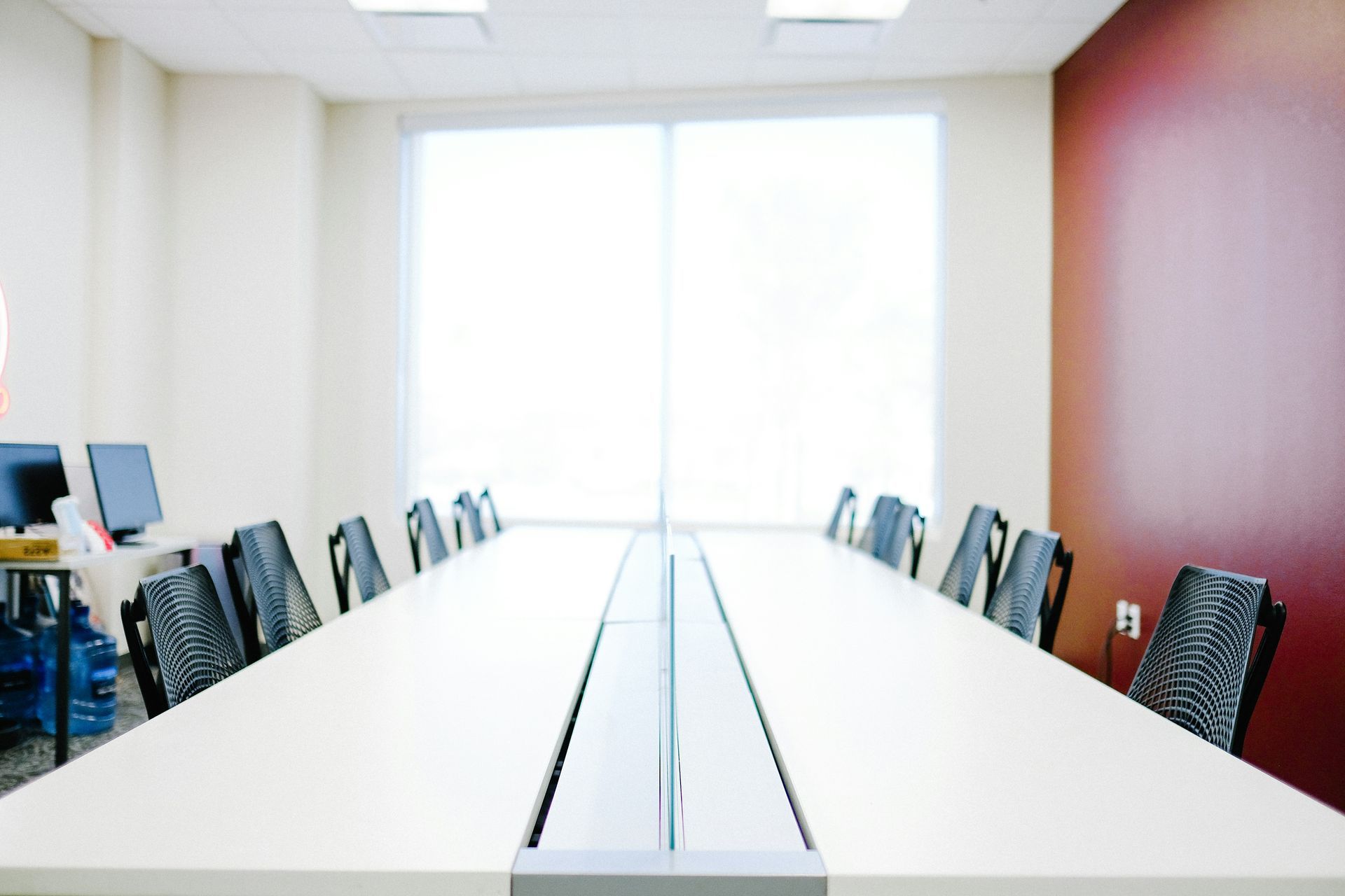 Long conference table in an empty room with black chairs, a large window, and a maroon accent wall.