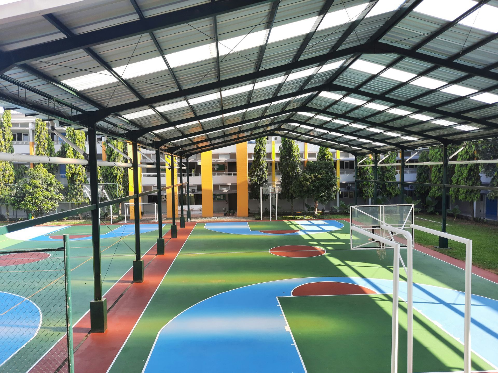 Covered outdoor multi-sport court with green, blue, and red surfaces; metal supports and roof.