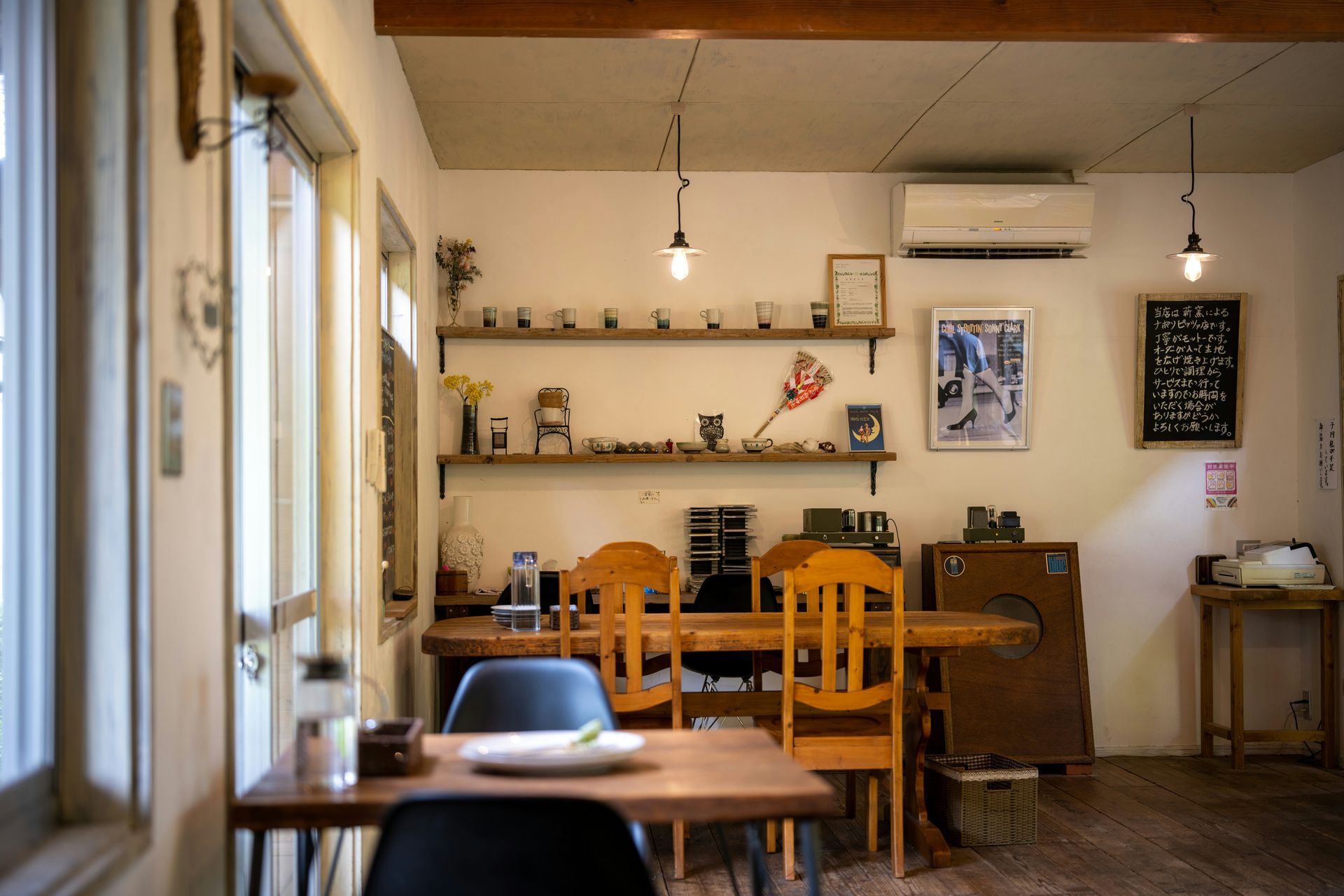 Interior view of a cafe with wooden tables, chairs, shelves, and decor. Sunlight streams in from a window.