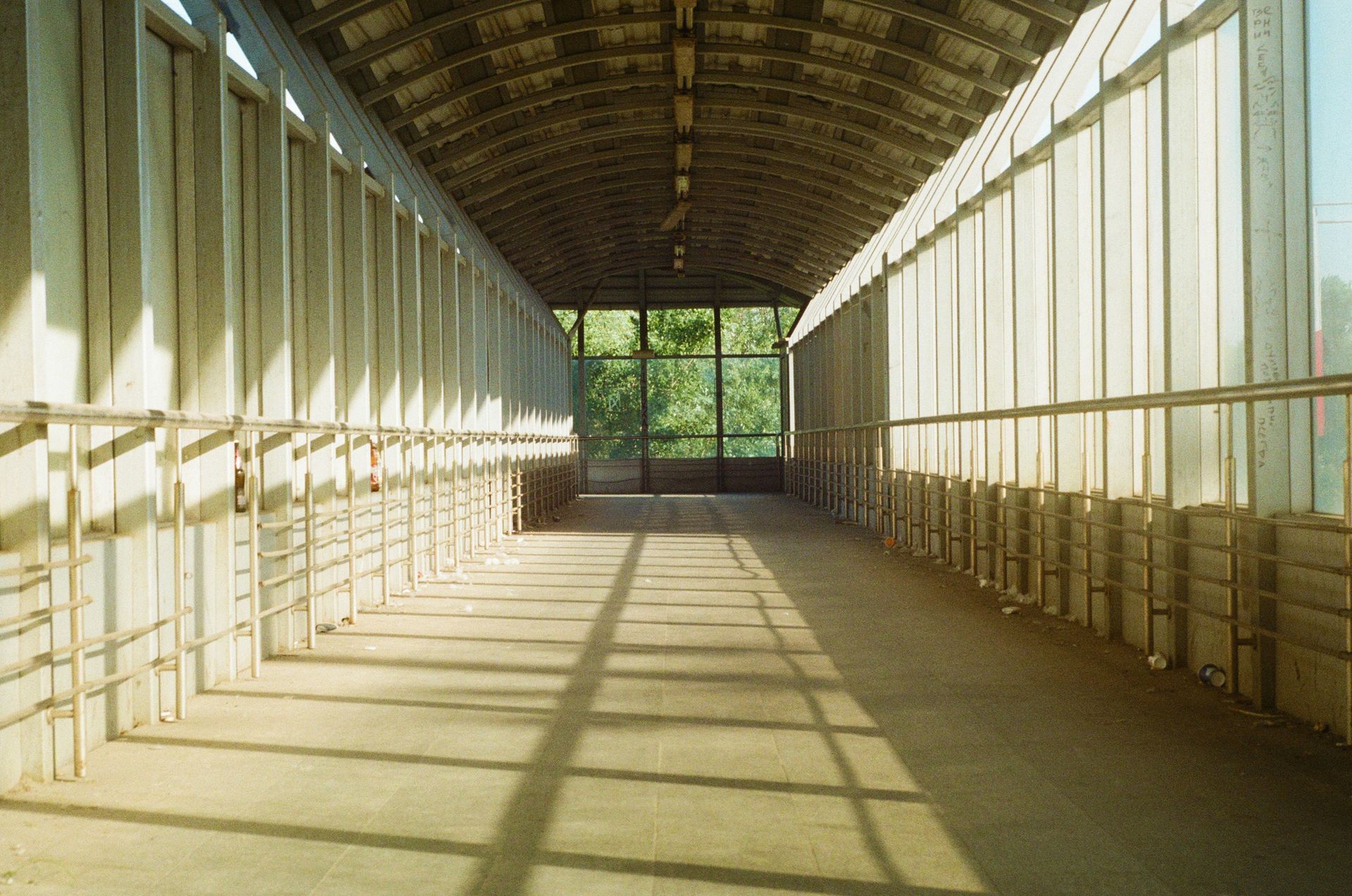 Covered walkway, arched ceiling, sunlight casts shadows on floor, leading to trees.