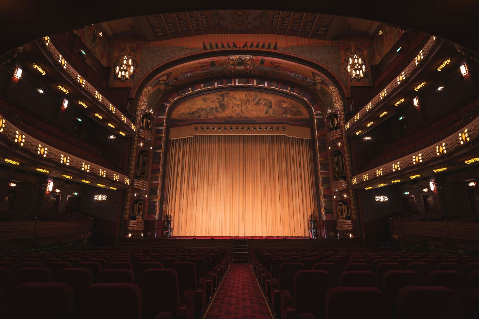 Inside an ornate, empty theater. Beige stage curtain, rows of red seats, and decorative walls.