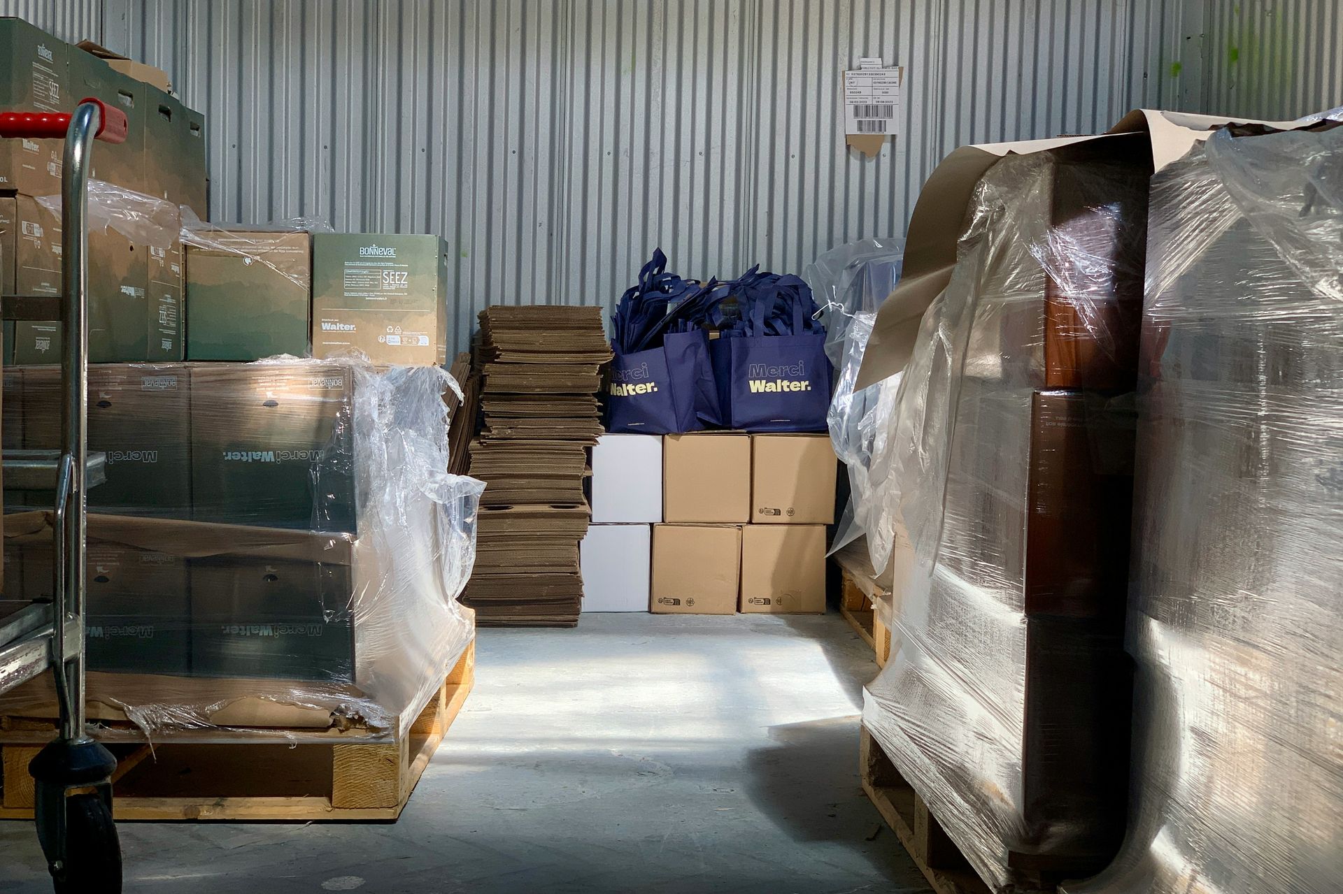 Warehouse interior with packed boxes on pallets, wrapped in plastic. A hand truck is visible on the left.