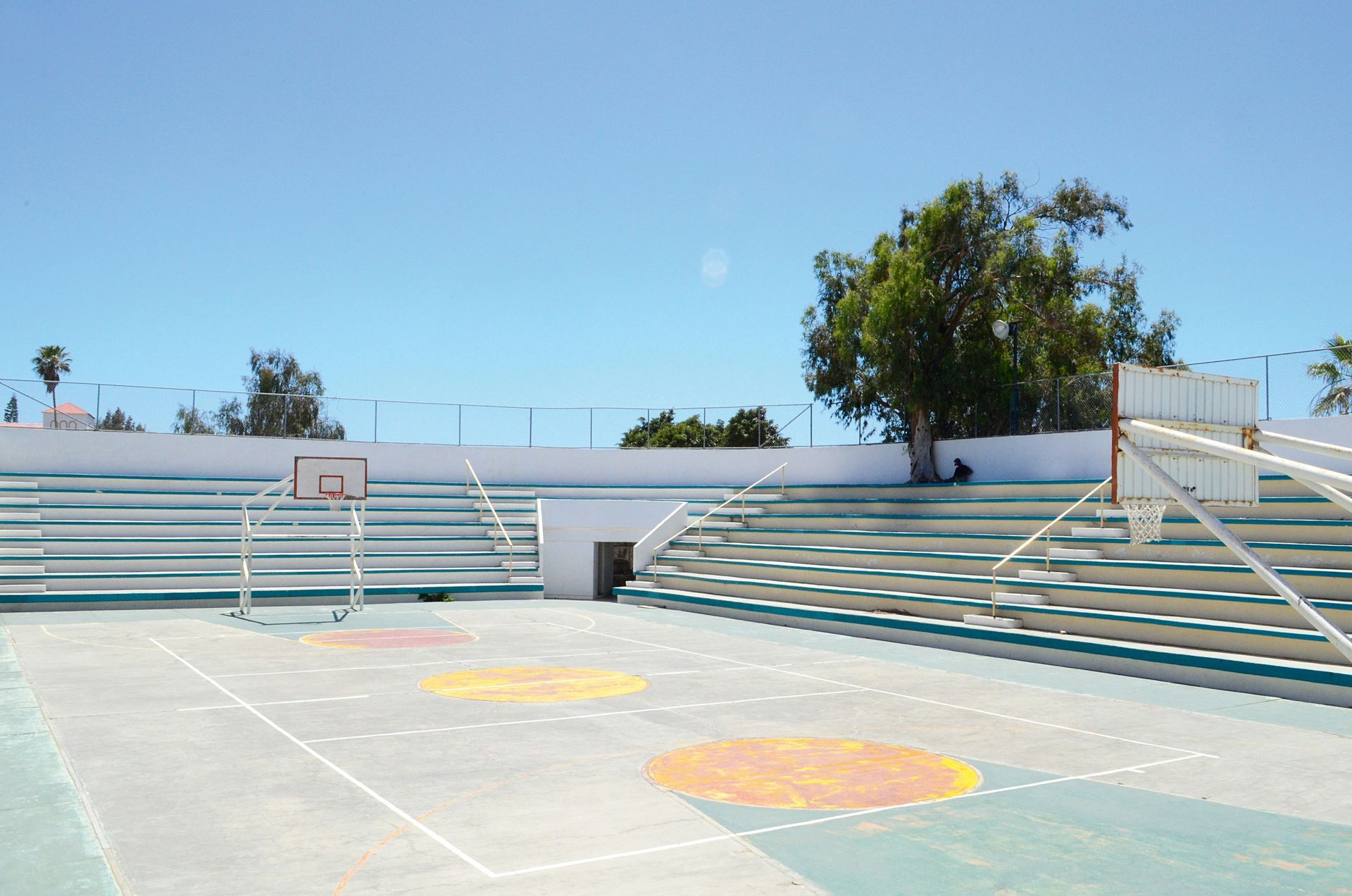 Outdoor sports court with basketball hoops, bleachers, and a tree under a bright blue sky.