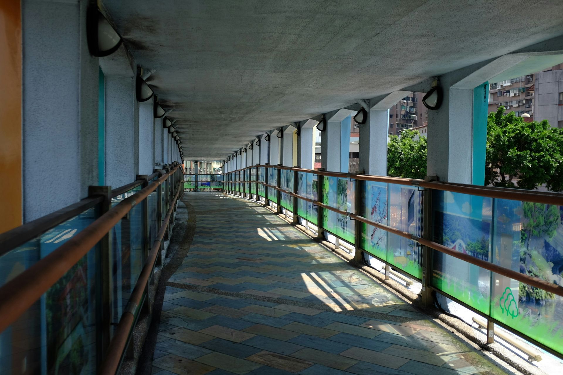 Covered walkway with glass panels and metal railings; exterior view of a city.