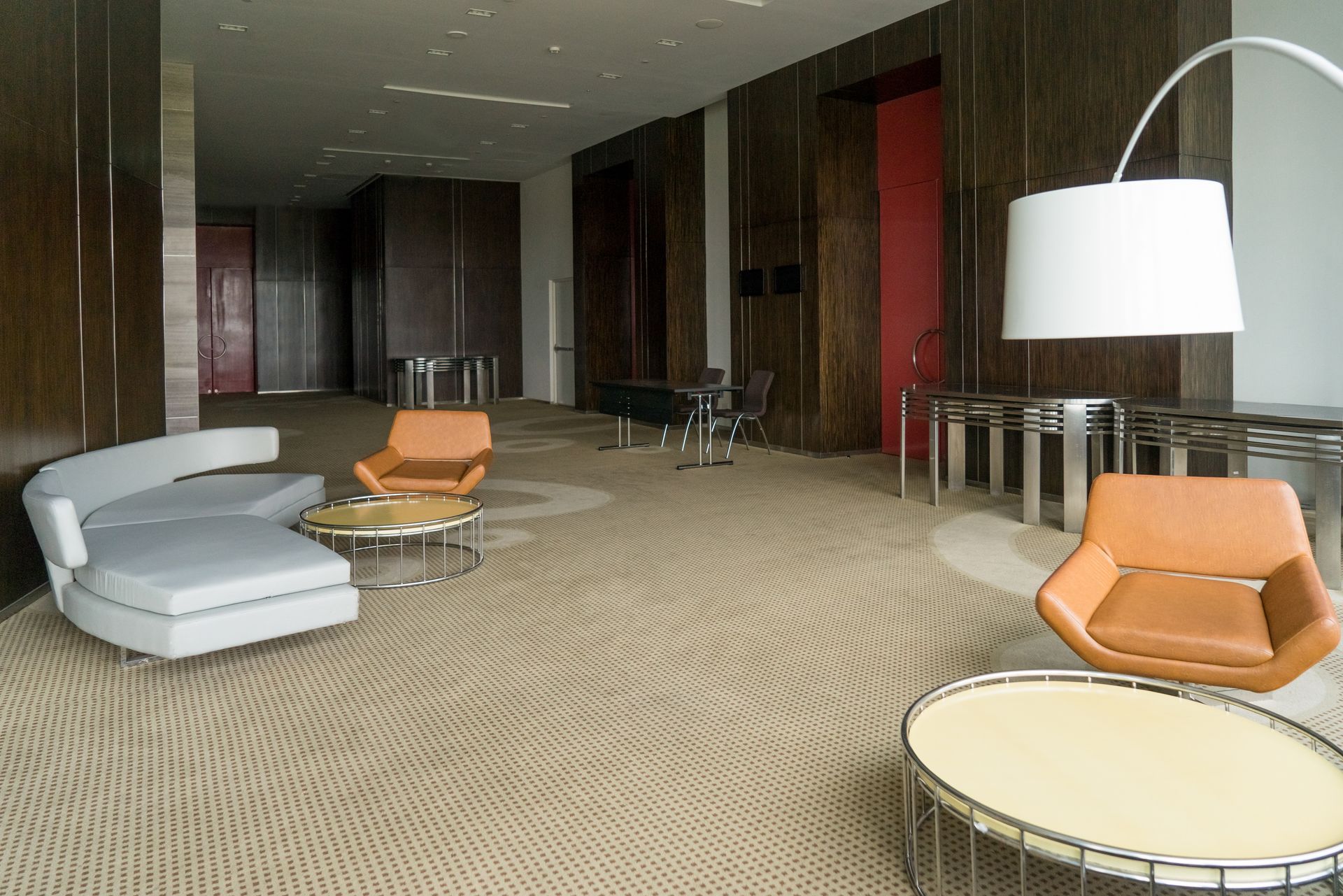 Modern lobby with seating, tables, and floor lamp. Brown walls and tan patterned carpet. Orange and gray furniture.