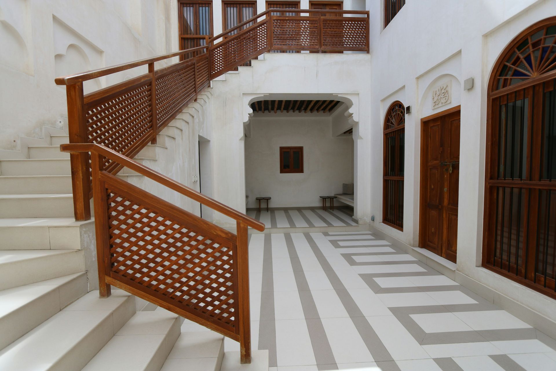 Courtyard with white walls, stairs with wooden railing, and patterned floor, brown doors.