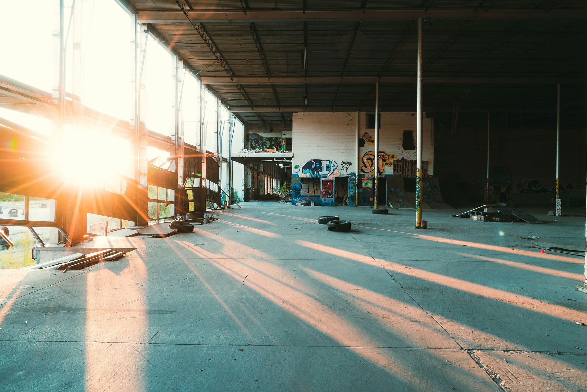 Sunlit abandoned warehouse, graffiti-covered walls, long shadows, debris on floor.
