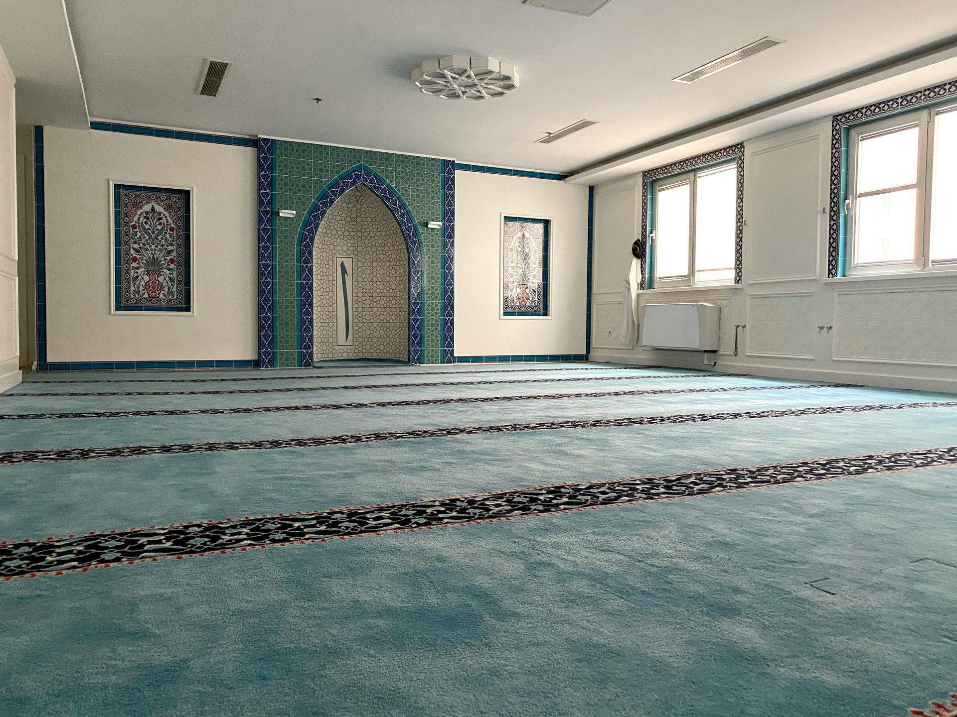 Inside of a mosque. Turquoise carpet with prayer lines, blue tile wall, and light from windows.