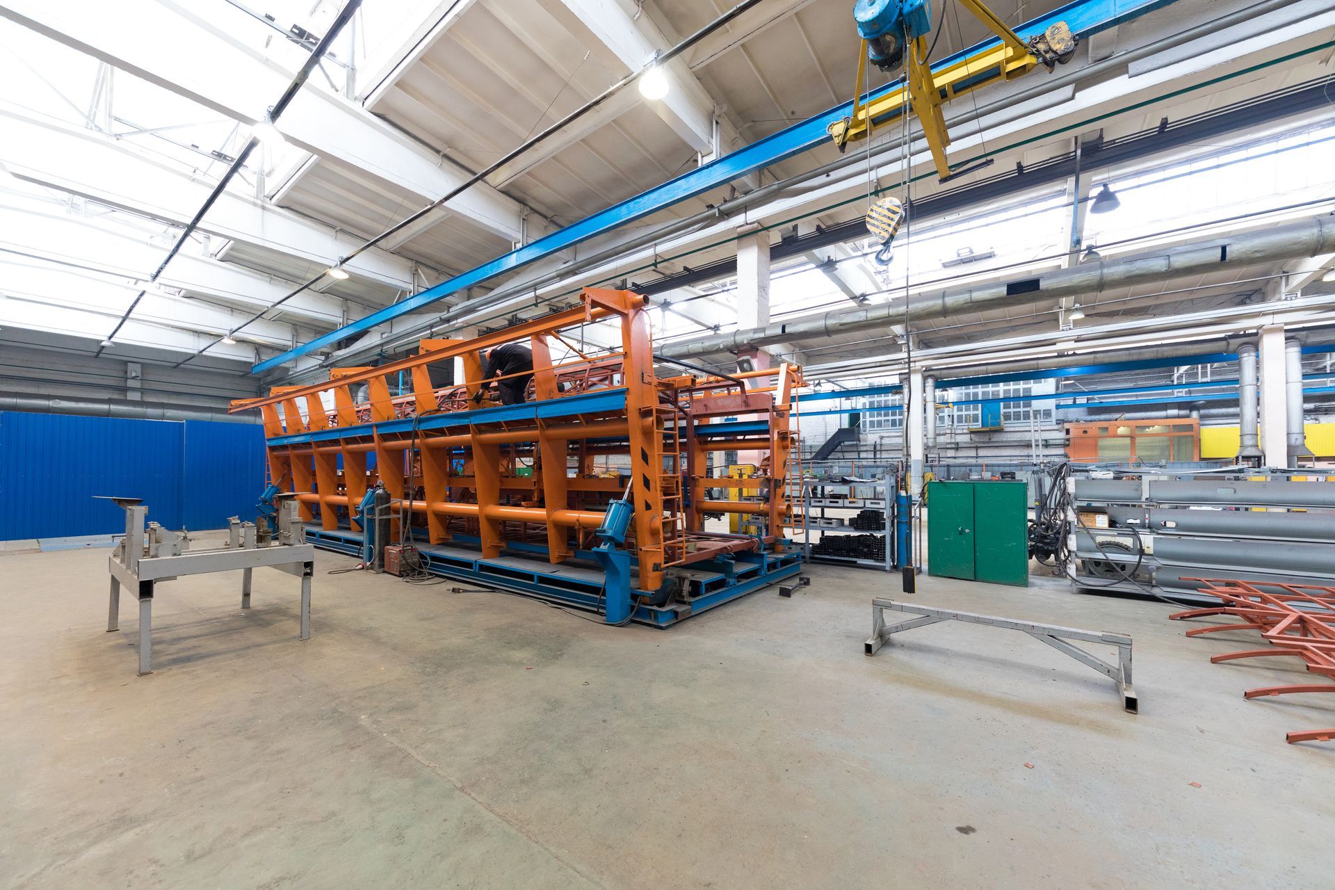 Orange and blue industrial machinery in a factory setting with overhead crane and metal materials.