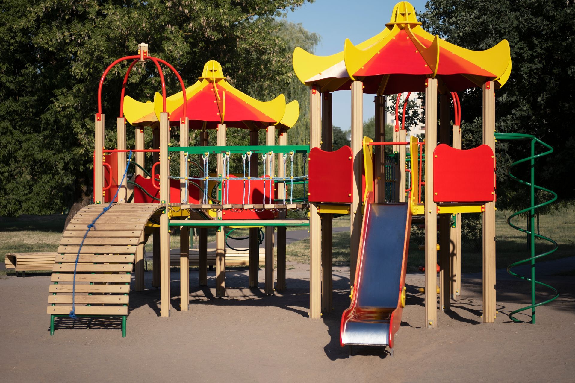 Playground with red, yellow, and green structures, including slides and climbing areas, set in a park.