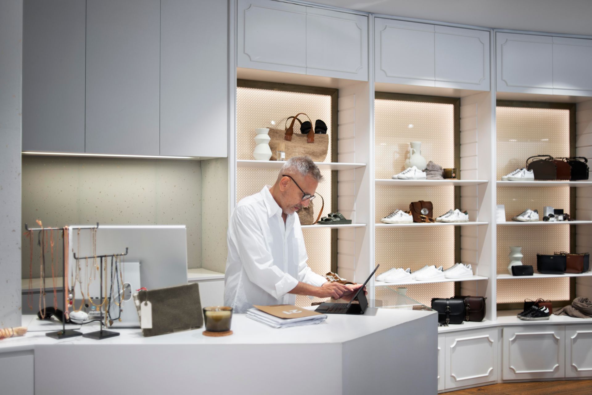 Man at a white counter in a boutique, working on a laptop. Shelves with shoes and bags in the background.