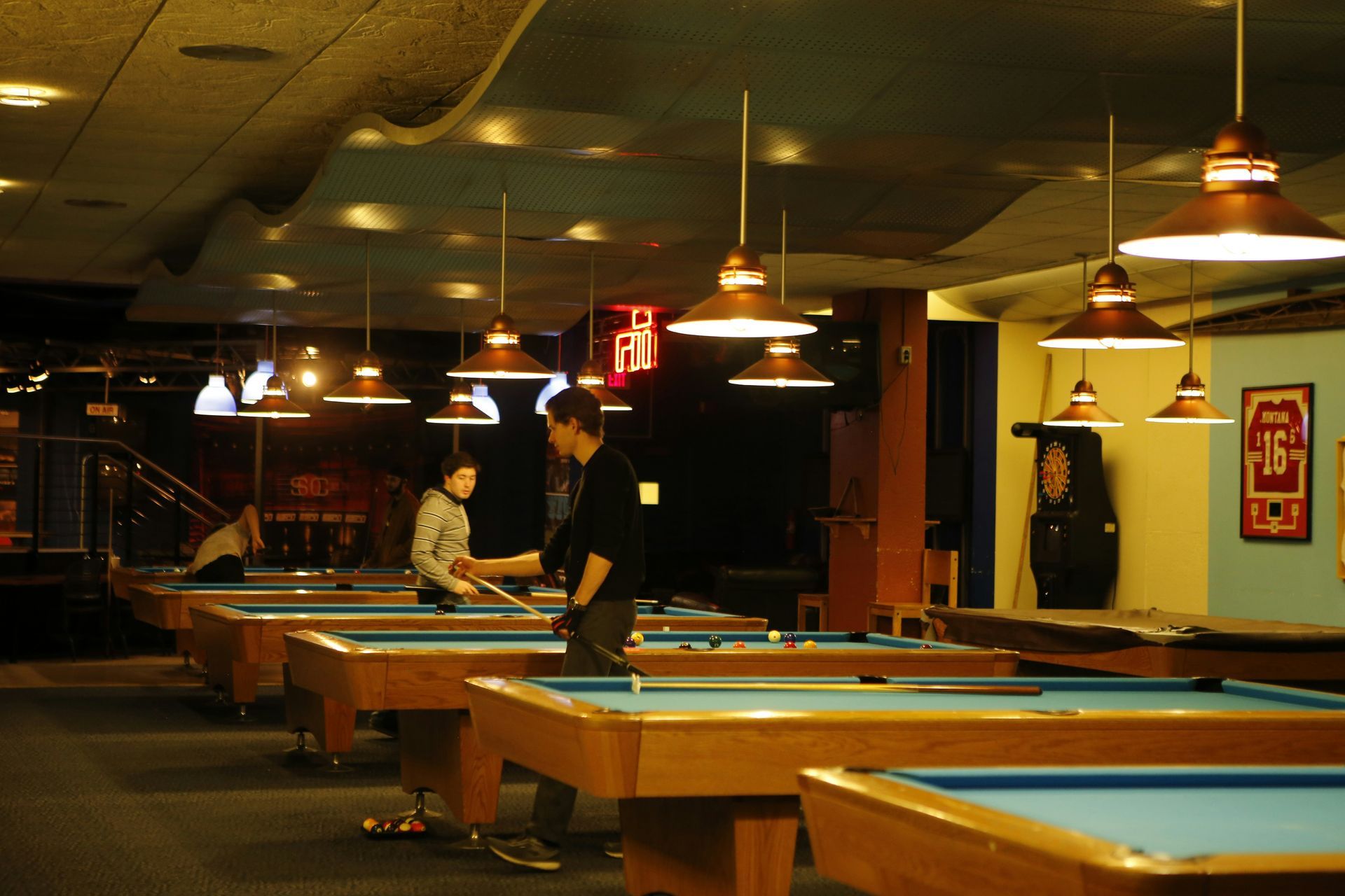 Pool hall with several tables, lit by hanging lamps. People play billiards.