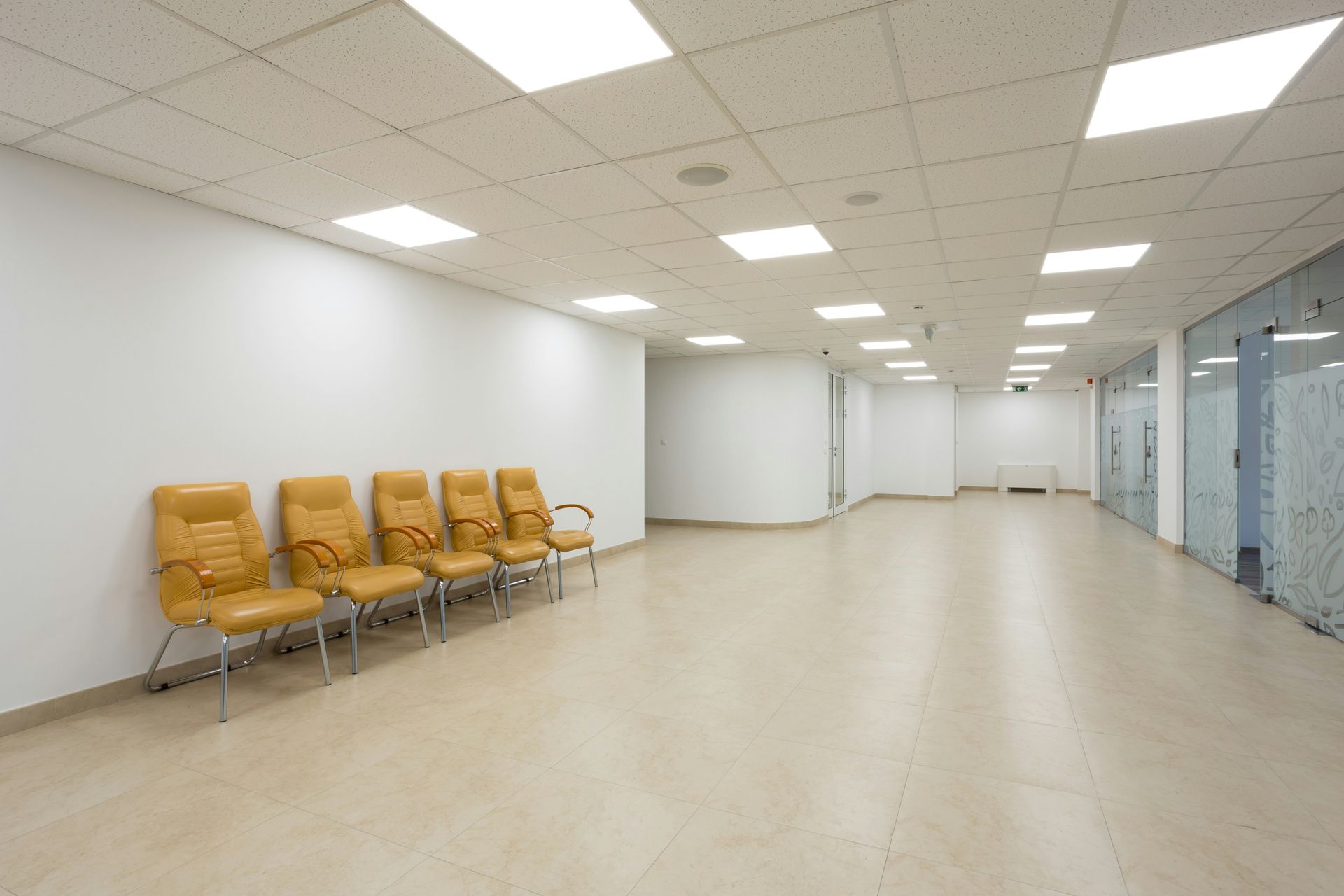 Waiting room with tan chairs against a white wall; beige floor and square ceiling lights.