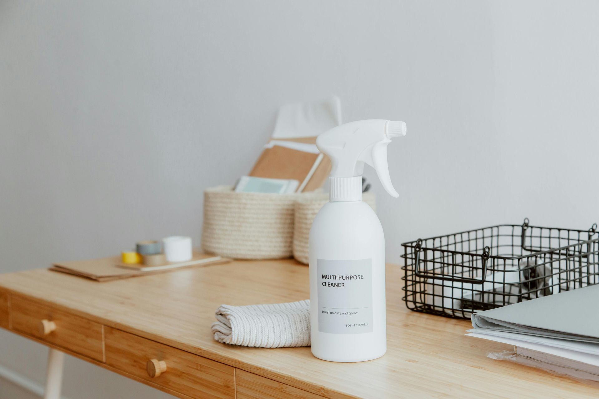 Spray bottle and cloth on a wooden desk with office supplies in the background.