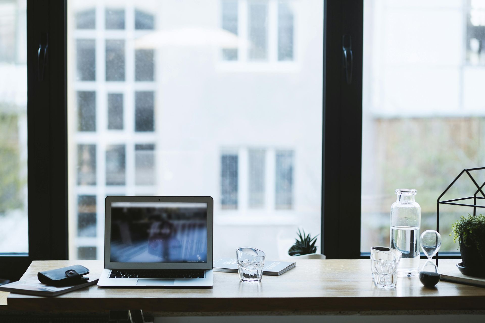 Laptop open on a wooden desk by a window; building view. Bottle and glass, small plant.