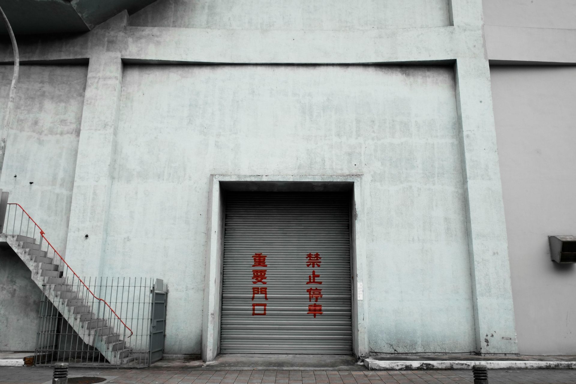 Gray concrete building with a recessed doorway. A metal roll-up door is partially open, displaying red Chinese text. Exterior stairs on the left.
