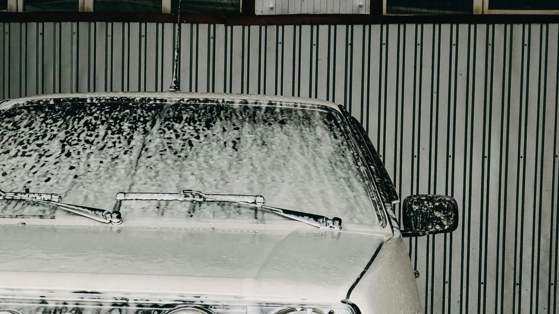 A white vehicle in Treasure Valley covered in thick foam cleaned well by Noble Clean in front of a metal wall; the windshield wipers are visible.