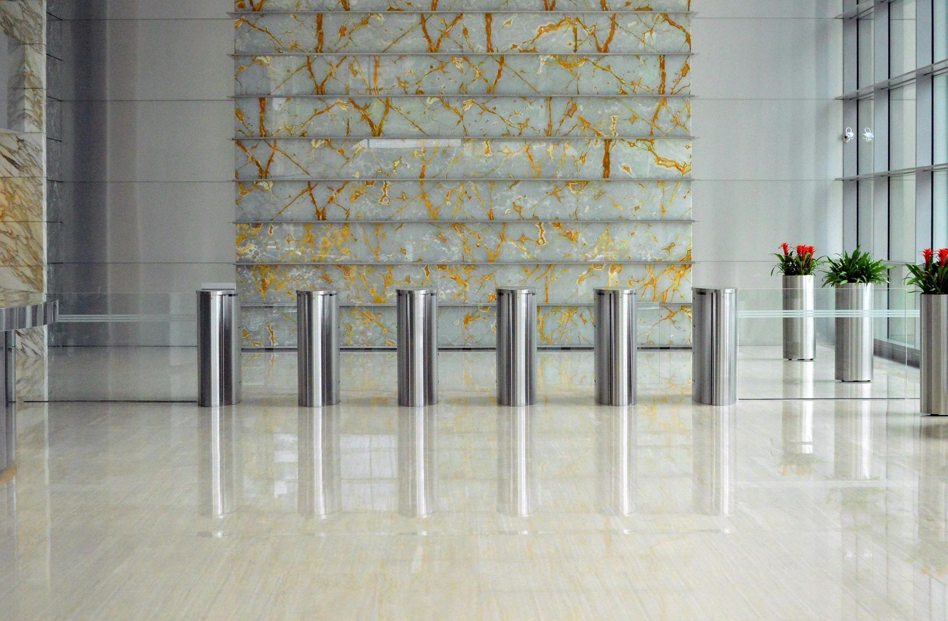 Stainless steel turnstiles in a polished lobby, reflecting overhead light. Marble wall backdrop, with potted plants nearby.