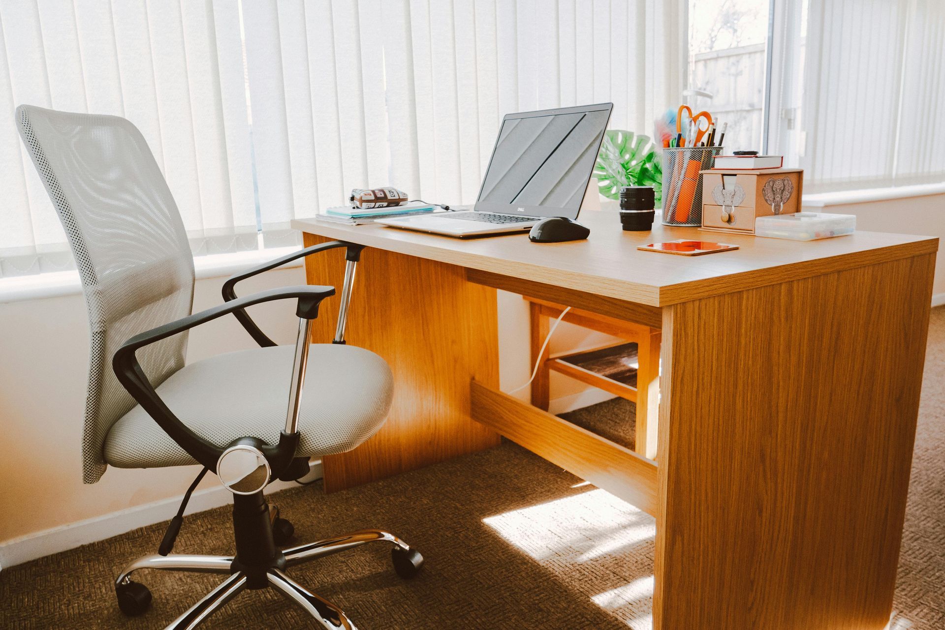 Office desk with a laptop, supplies, and an office chair near a window.