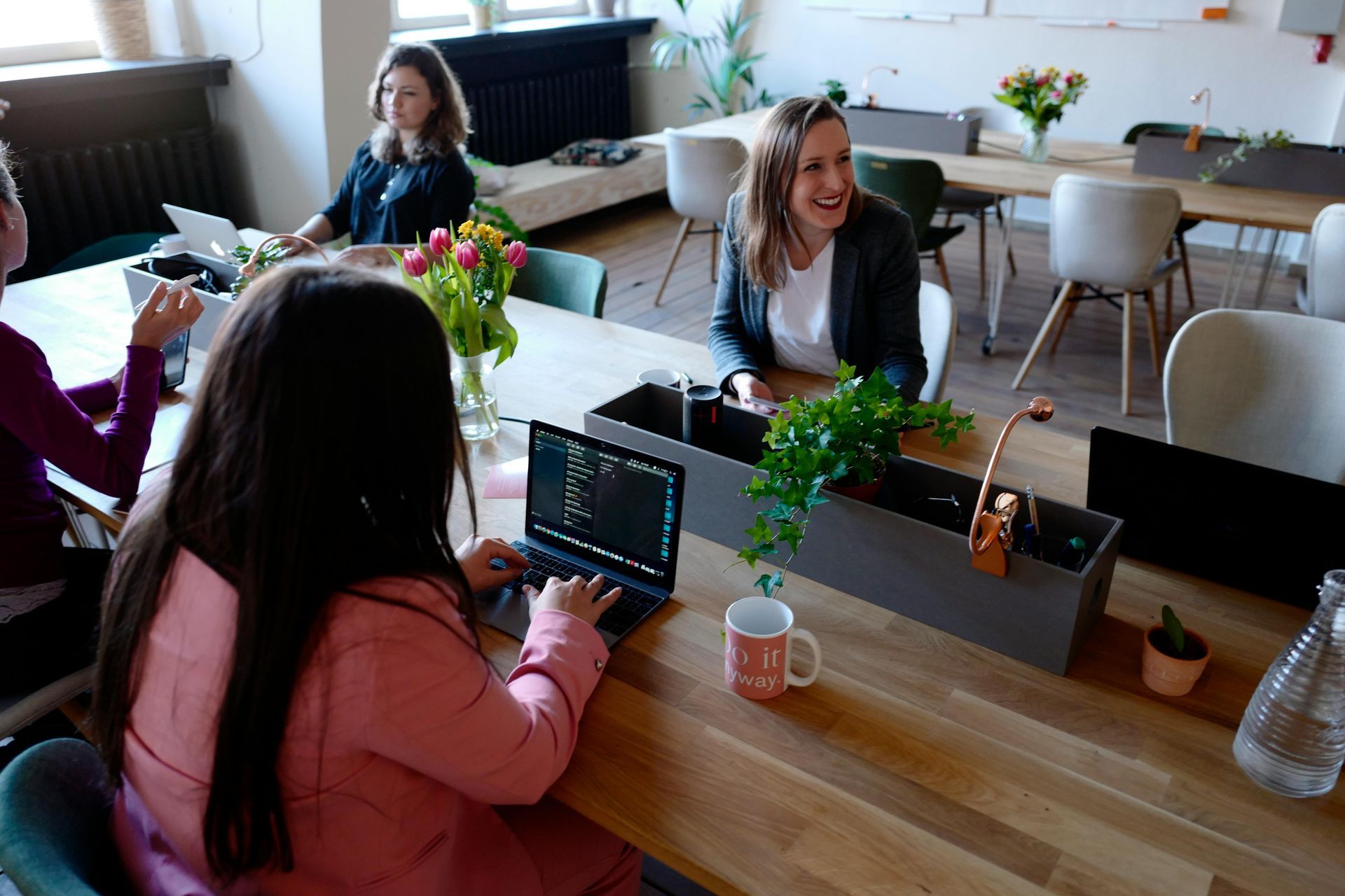 Four people in an office setting around a wooden table. One woman types on a laptop, others smile, and flowers are present.