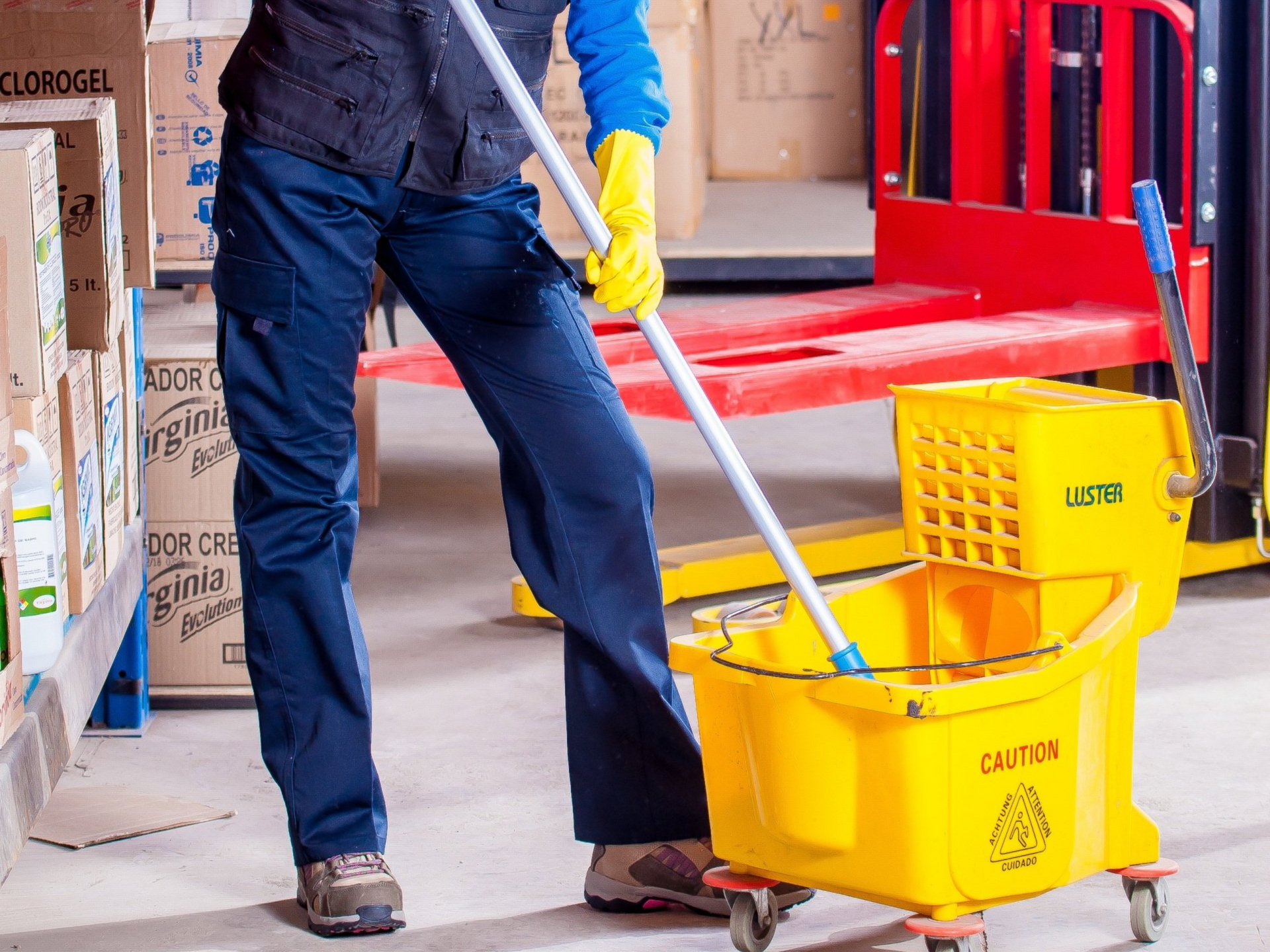 A person wearing yellow cleaning gloves mops the floor next to a yellow industrial mop bucket in a warehouse.