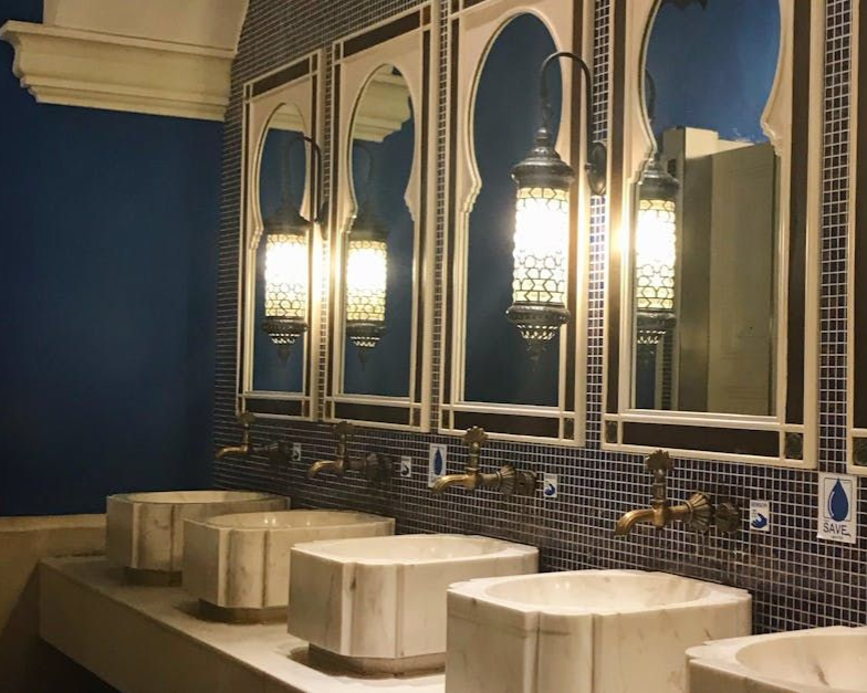 Bathroom with blue tile, arched mirrors, ornate lamps, and stone sinks.