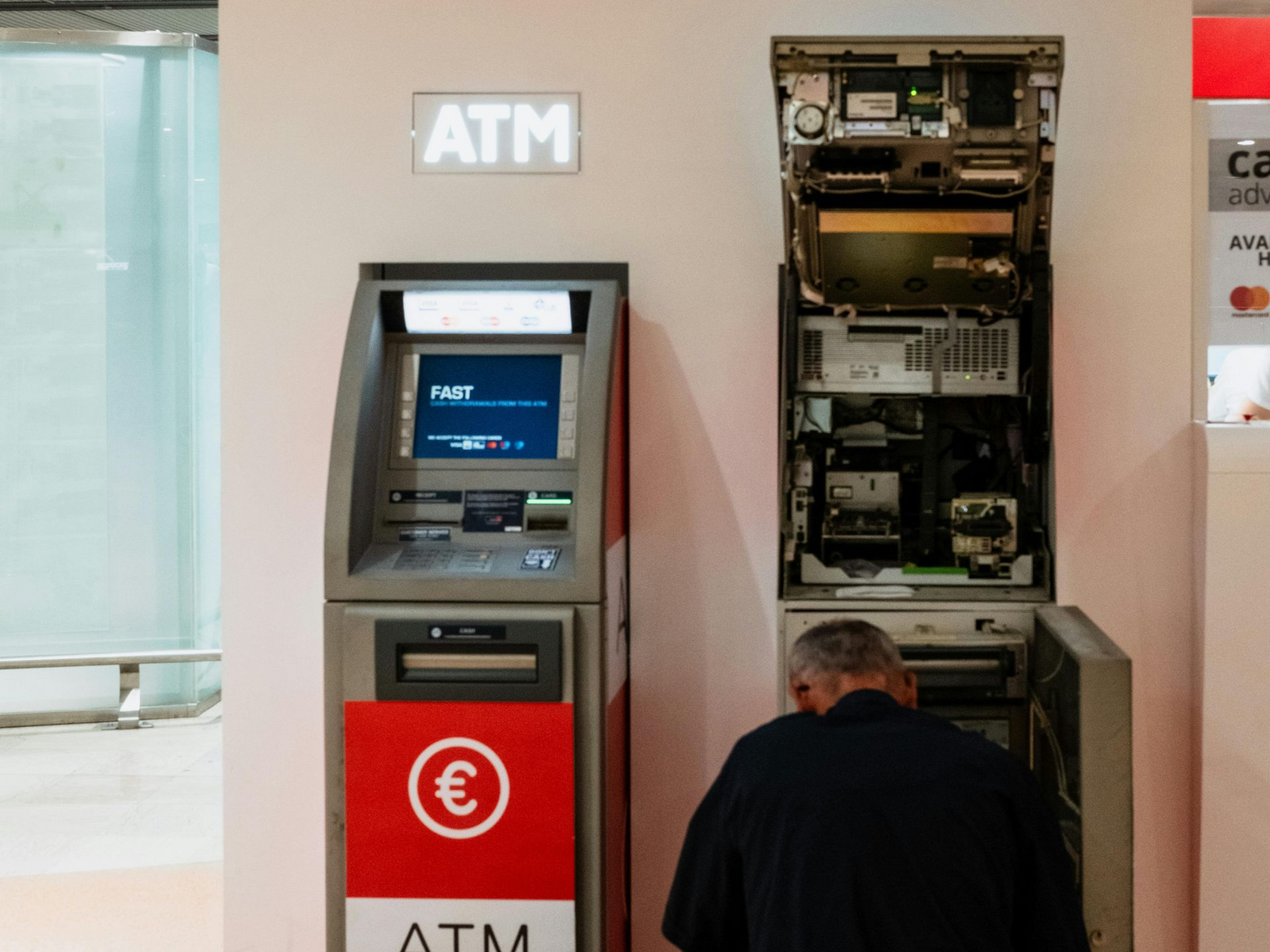 Man repairing ATM in a Nampa bank, with the machine's interior exposed. Red ATM signage is visible.